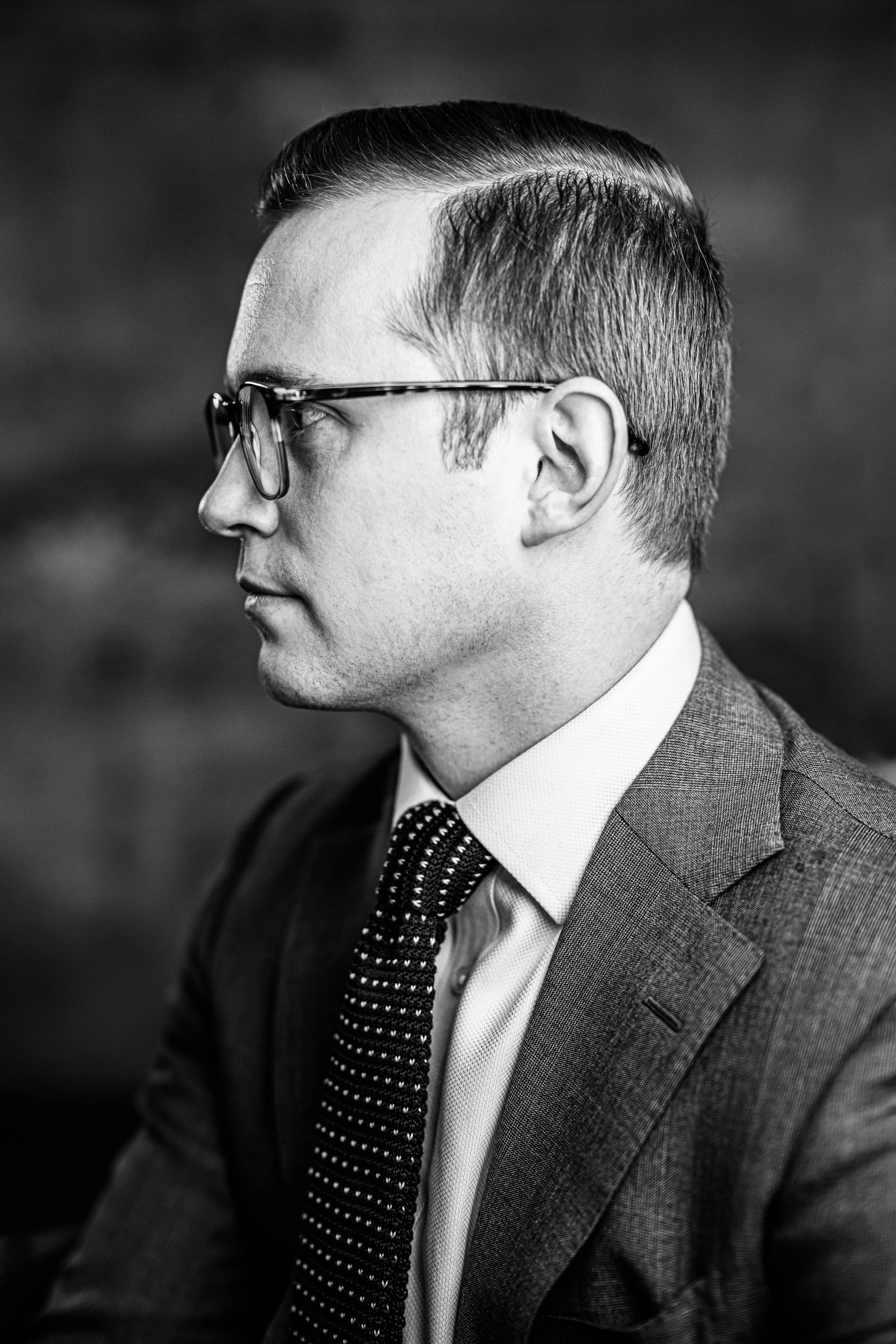 Black and white portrait of a man in glasses, dressed in a suit and tie, in profile view.