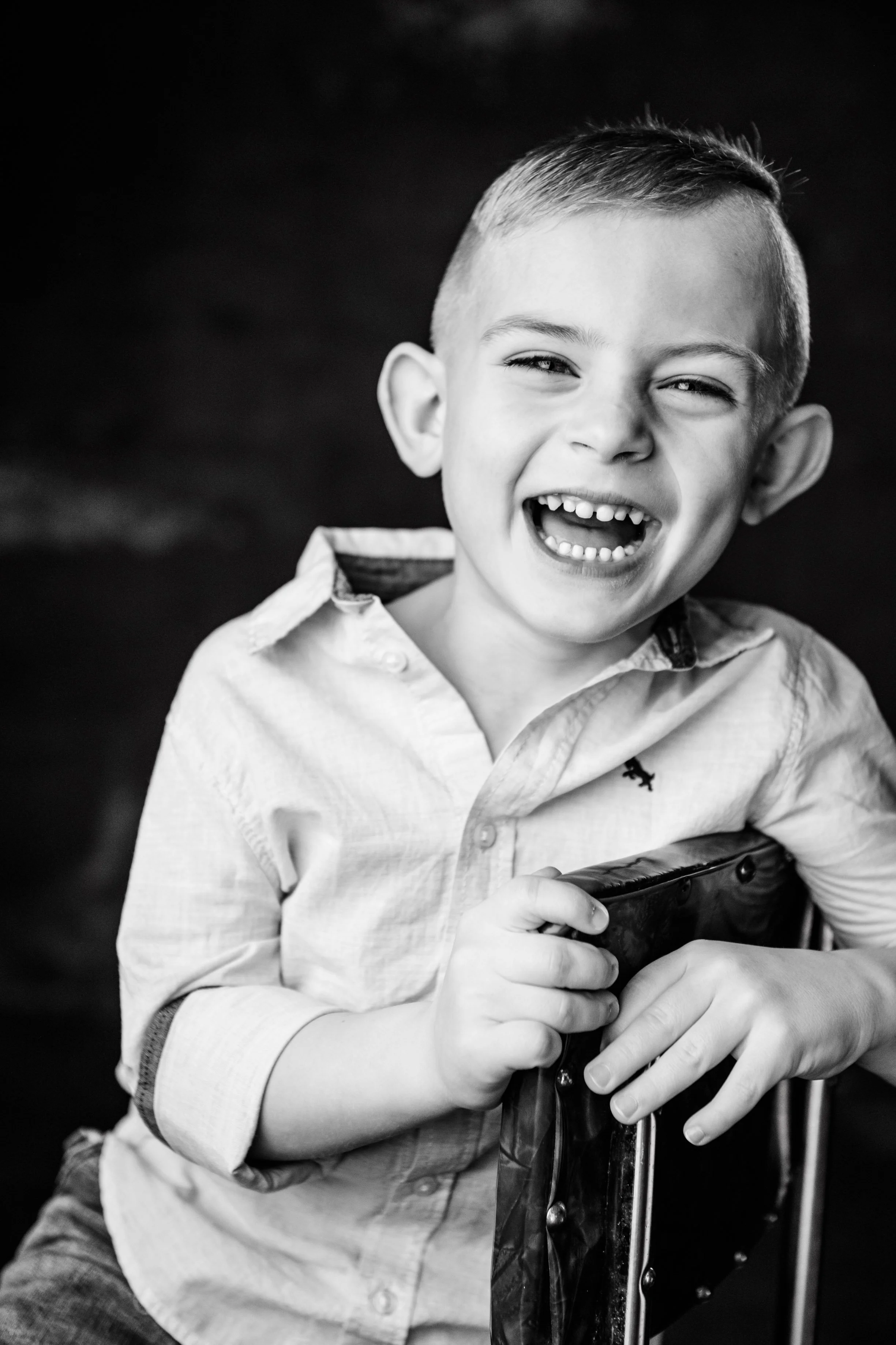 A smiling young boy with short hair, wearing a light-colored button-up shirt, holding onto a piece of furniture, possibly a small chair or stool, with his left hand, laughing or in a moment of joy, in black and white.
