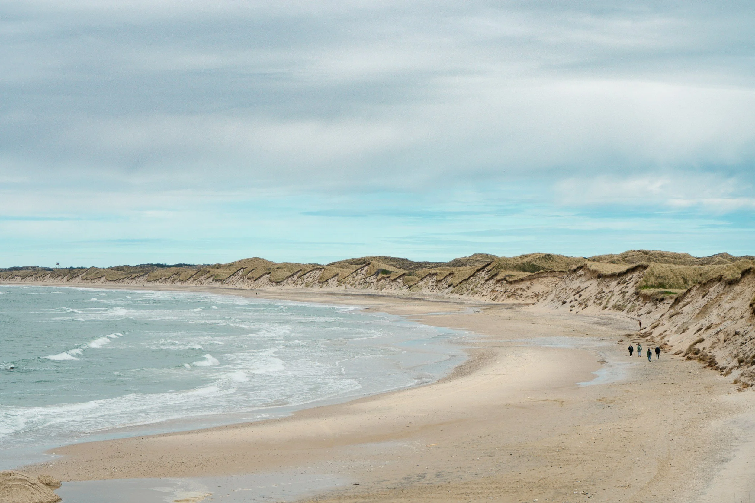 A sandy beach with greenish-blue ocean water lapping against the shore. Dunes with grass line the top of the beach, and a few people are walking along the shoreline under a cloudy sky.