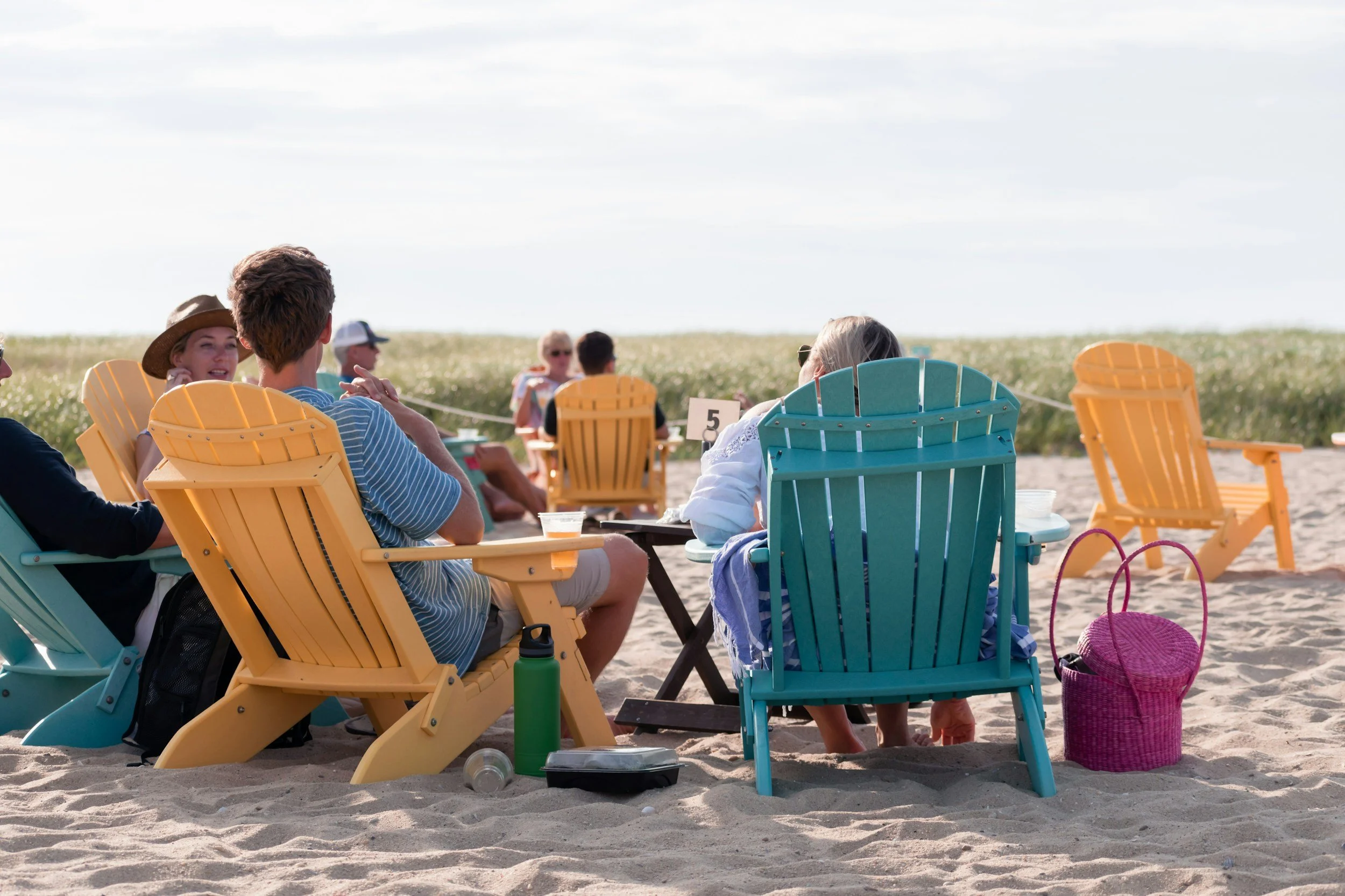 People sitting on colorful Adirondack chairs on a sandy beach, engaged in conversation, with a clear sky and distant greenery in the background.