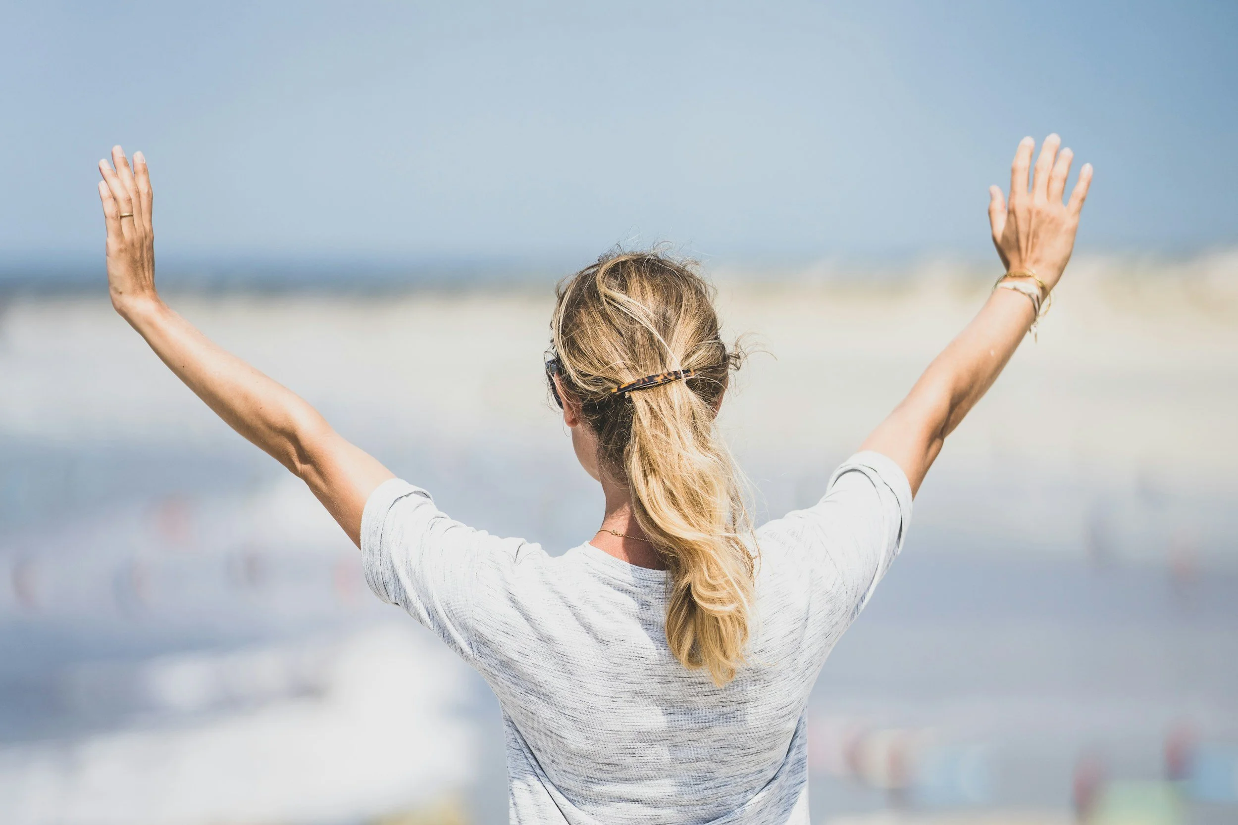 A woman with long blonde hair in a ponytail wearing sunglasses and a white t-shirt, standing on a beach with her arms raised and facing the ocean.