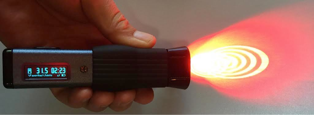 Hand holding a handheld device emitting a red laser beam with concentric ripples on a gray surface.