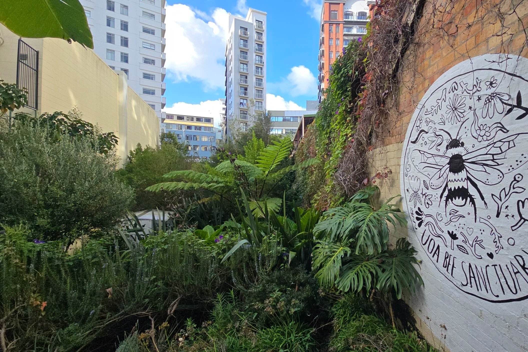 Urban garden with lush green plants, trees, and shrubs, alongside a brick wall with a white mural featuring a bee, flowers, and text, with modern high-rise buildings in the background under a partly cloudy sky.