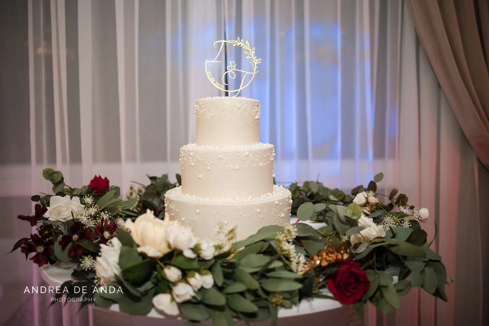 White three-tier wedding cake decorated with pearls, topped with a gold cake topper, surrounded by a ring of white and red roses and greenery.