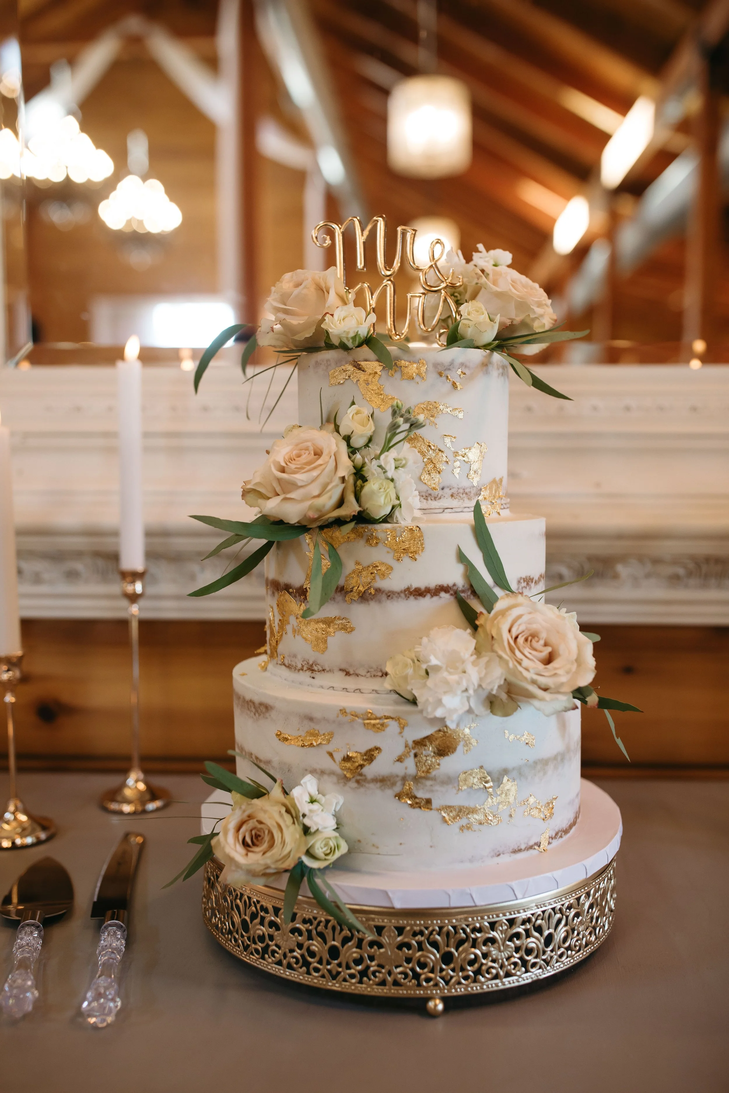 A three-tiered wedding cake with white and gold details decorated with cream and blush roses, along with greenery. The cake sits on an ornate gold cake stand in a rustic venue with wooden beams and hanging lights.