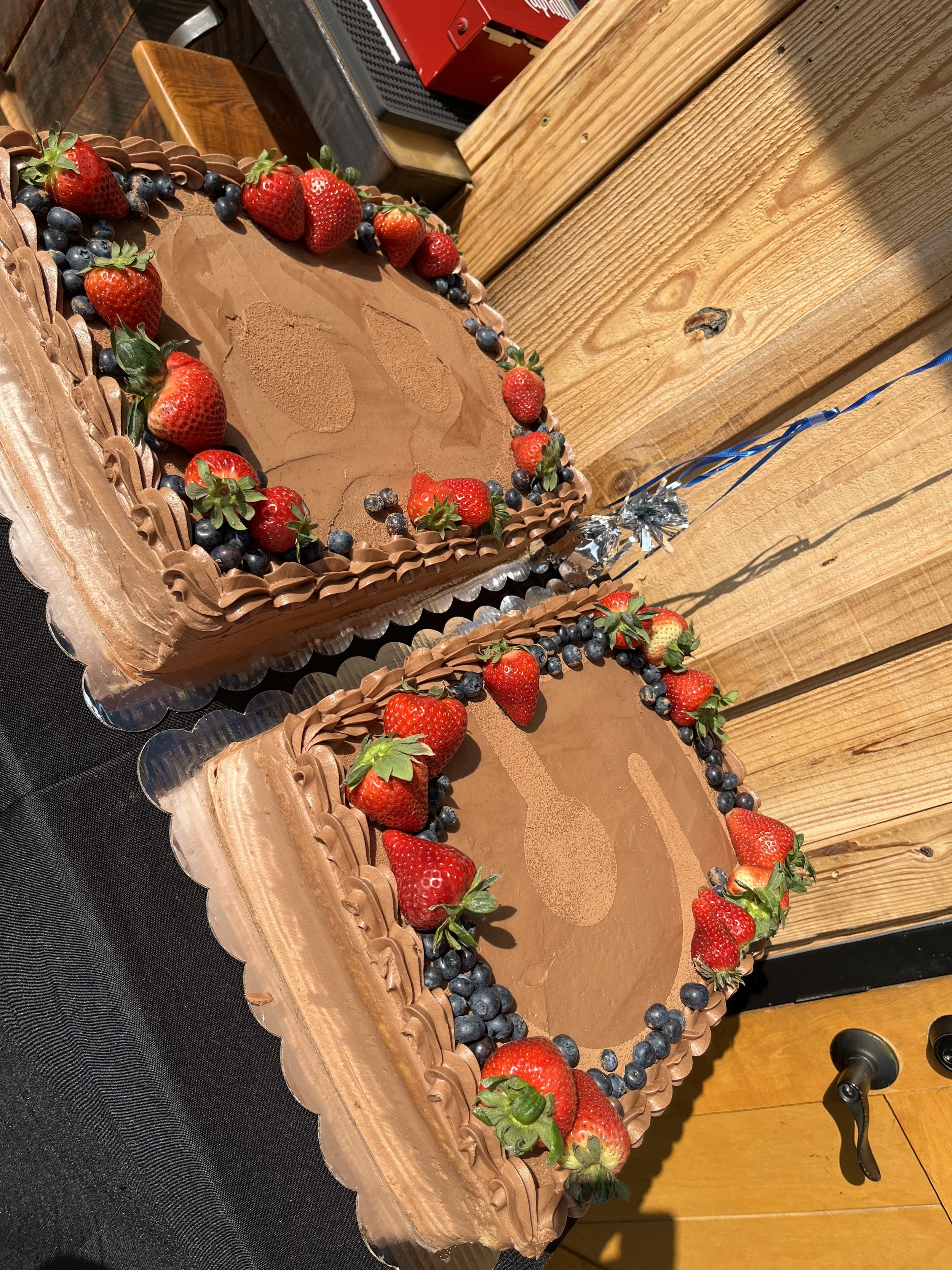 Two rectangular chocolate cakes decorated with strawberries and blueberries, with a cupcake shape pattern in the center of each cake, placed on a black table next to a wooden wall.