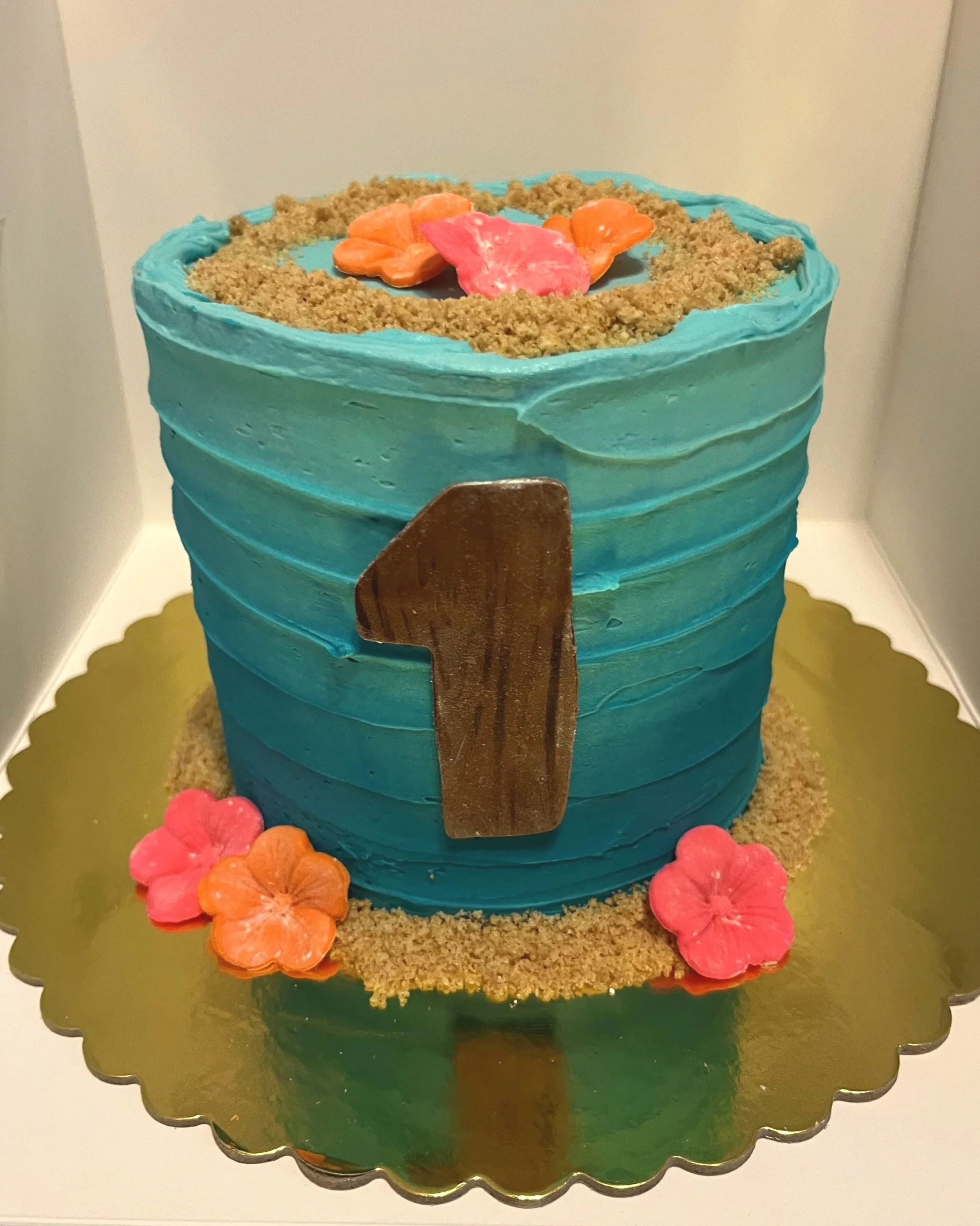 A blue birthday cake with a large gold number one on the front, decorated with pink and orange flowers, crumbly edible decorations on top and around the base, placed on a gold scalloped cake board.