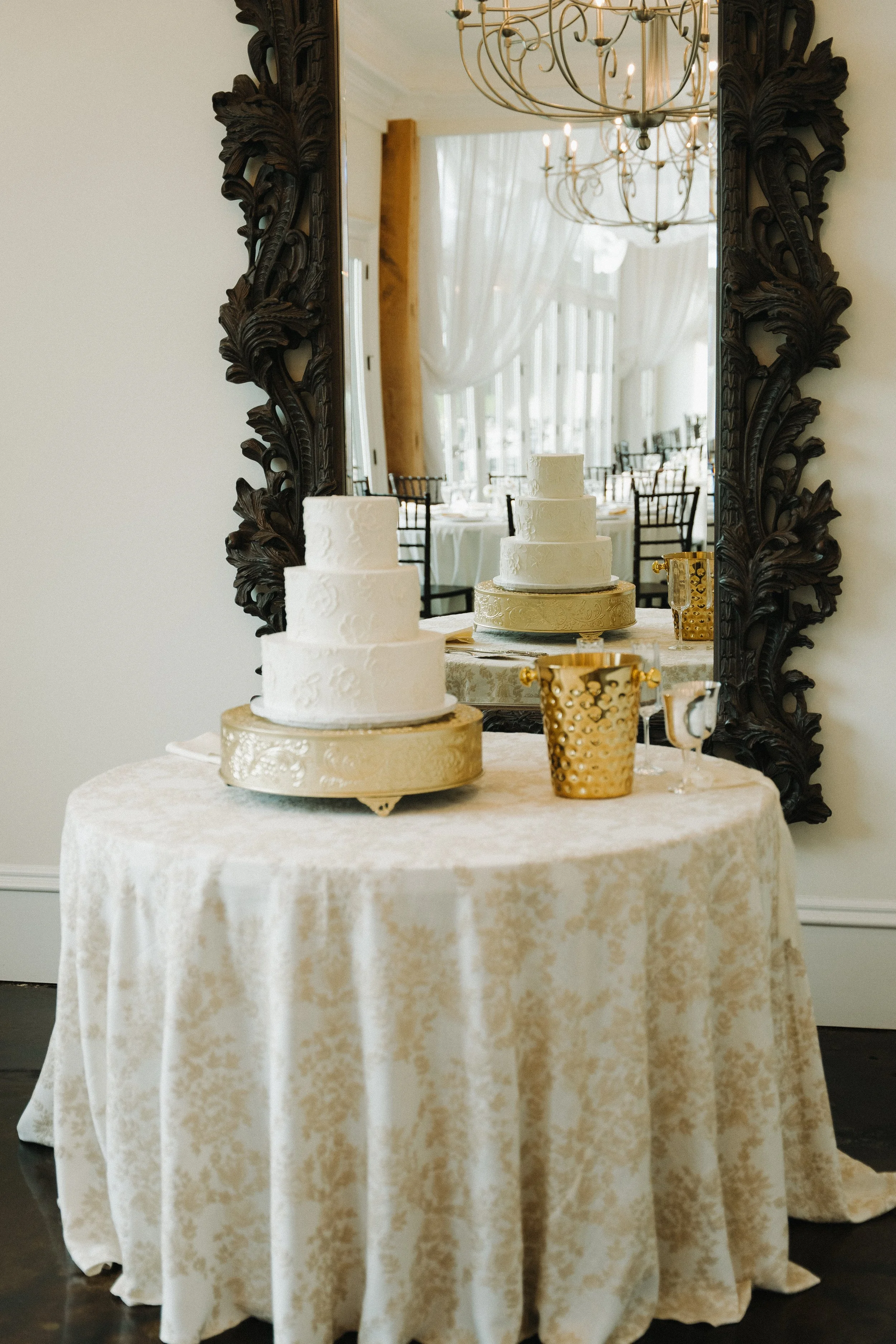 A round table covered with a patterned tablecloth, holding a tiered white wedding cake on a gold stand, gold decorative cups, and a partially seen gold-colored tray, with a large ornate mirror reflecting the cake and a chandelier in the background.