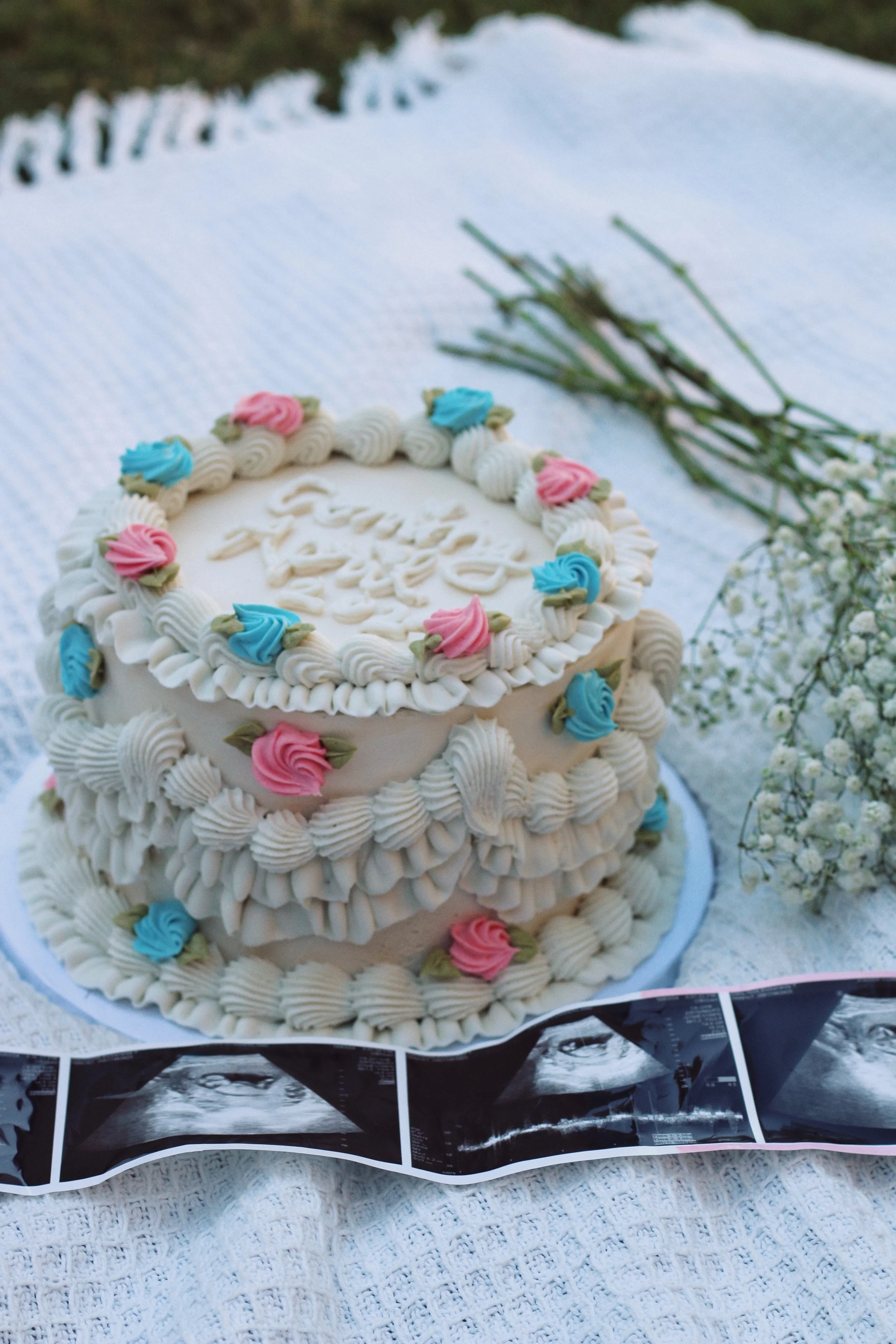 A white cake decorated with pink and blue roses, baby blue and white frosting, and an ultrasound image at the base, placed on a white textured cloth with a bouquet of white flowers nearby.