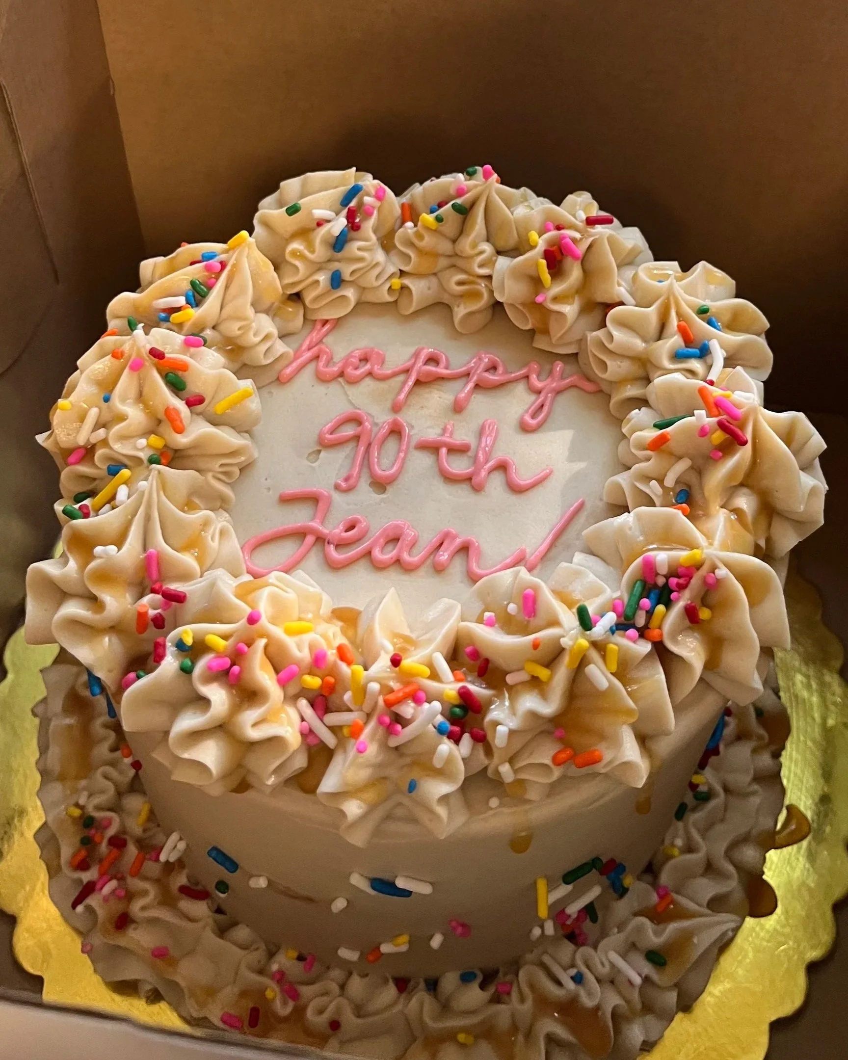 A round birthday cake with white frosting, decorated with swirls and colorful rainbow sprinkles. The top center has pink written message that says 'happy 90th Jean'.
