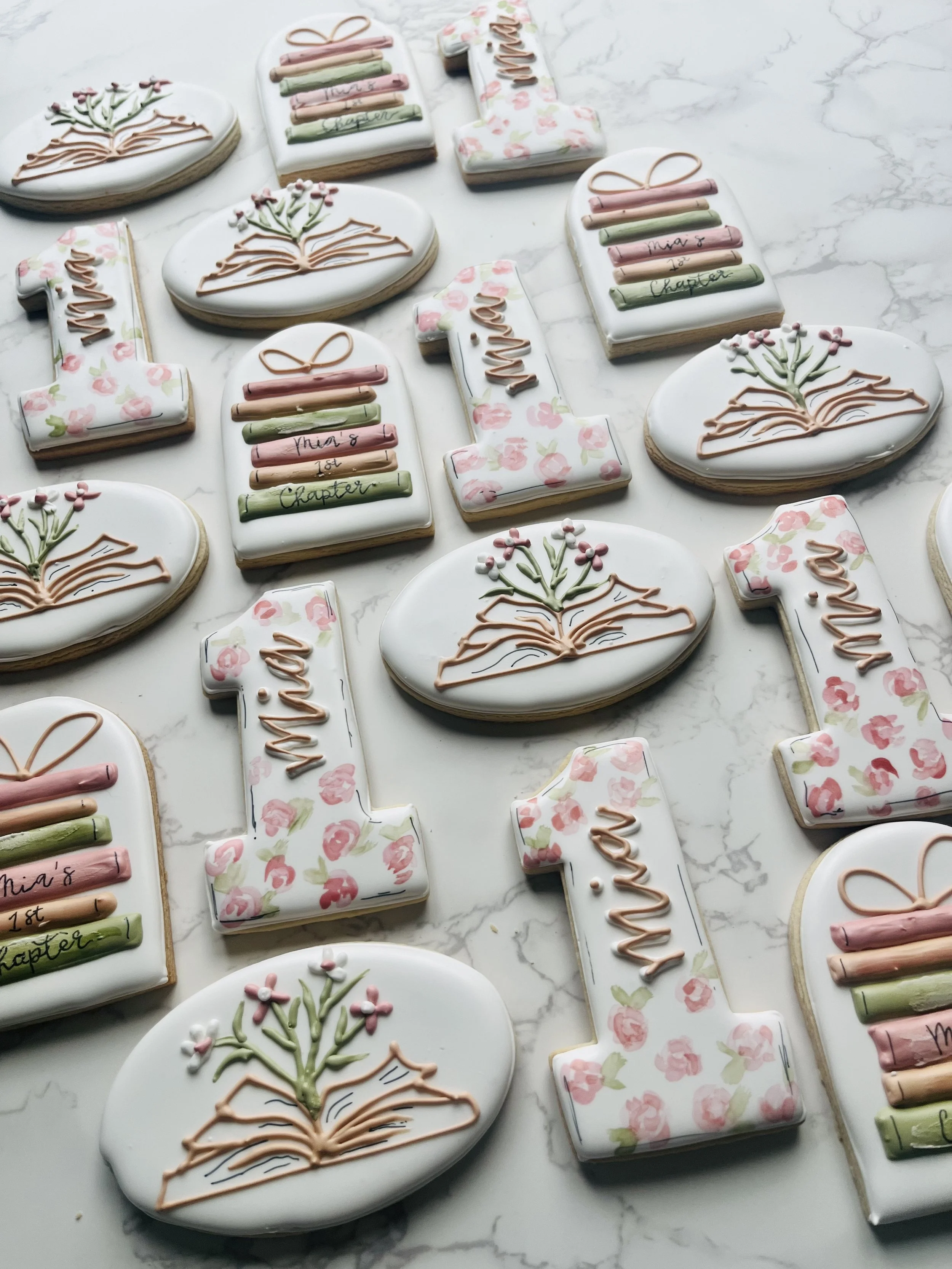 Decorated cookies on a white marble surface. The cookies are shaped as the number 1 and the letter L, with floral and book designs, and icing in pastel pink, green, and white colors.