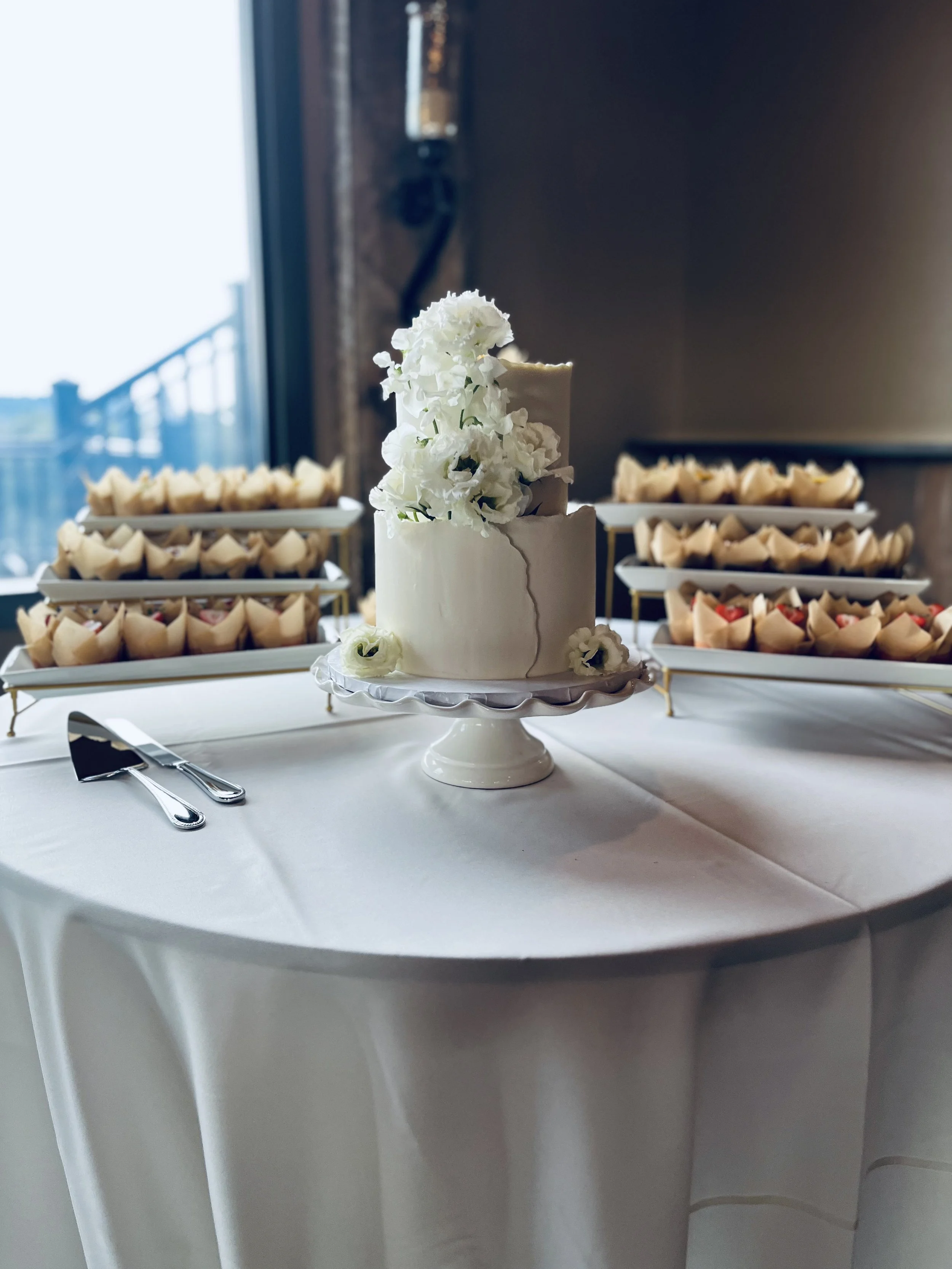A white wedding cake with white flowers, displayed on a cake stand in front of a table with assorted cupcakes on tiered trays.
