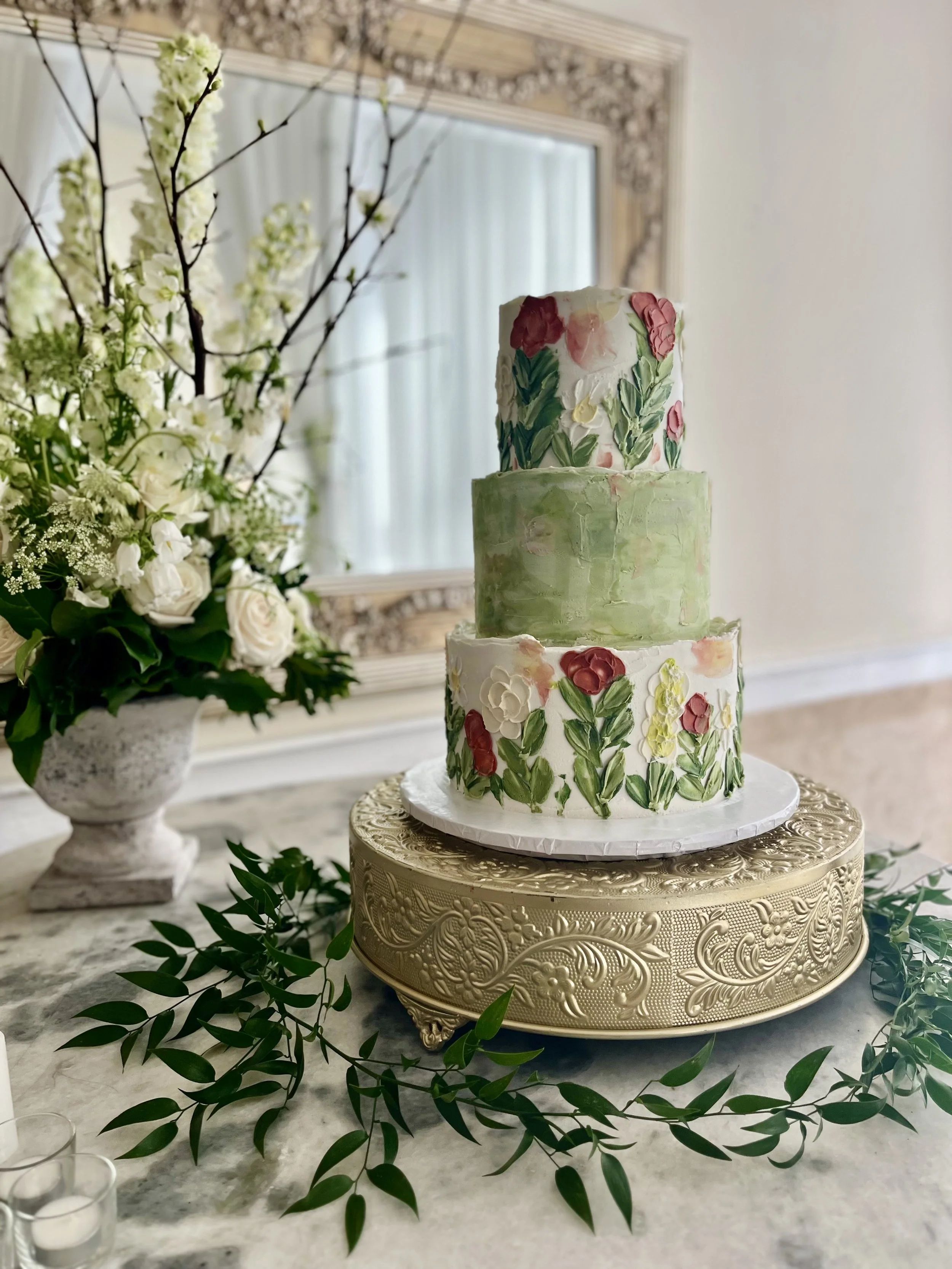 A three-tiered wedding cake with floral decorations, placed on an ornate gold cake stand. The cake features pastel green and white frosting with pink and white floral accents. A floral arrangement with white roses and greenery is visible in the background.