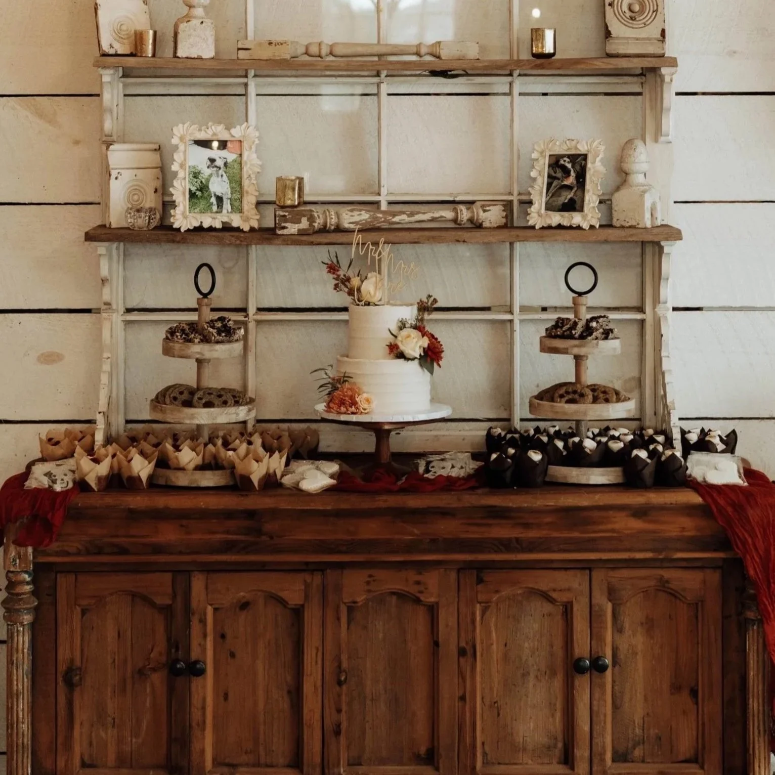 Decorative wedding dessert table featuring a white two-tiered cake with floral accents, surrounded by cookies, cupcakes, and framed photos on a rustic wooden and white shelving backdrop.