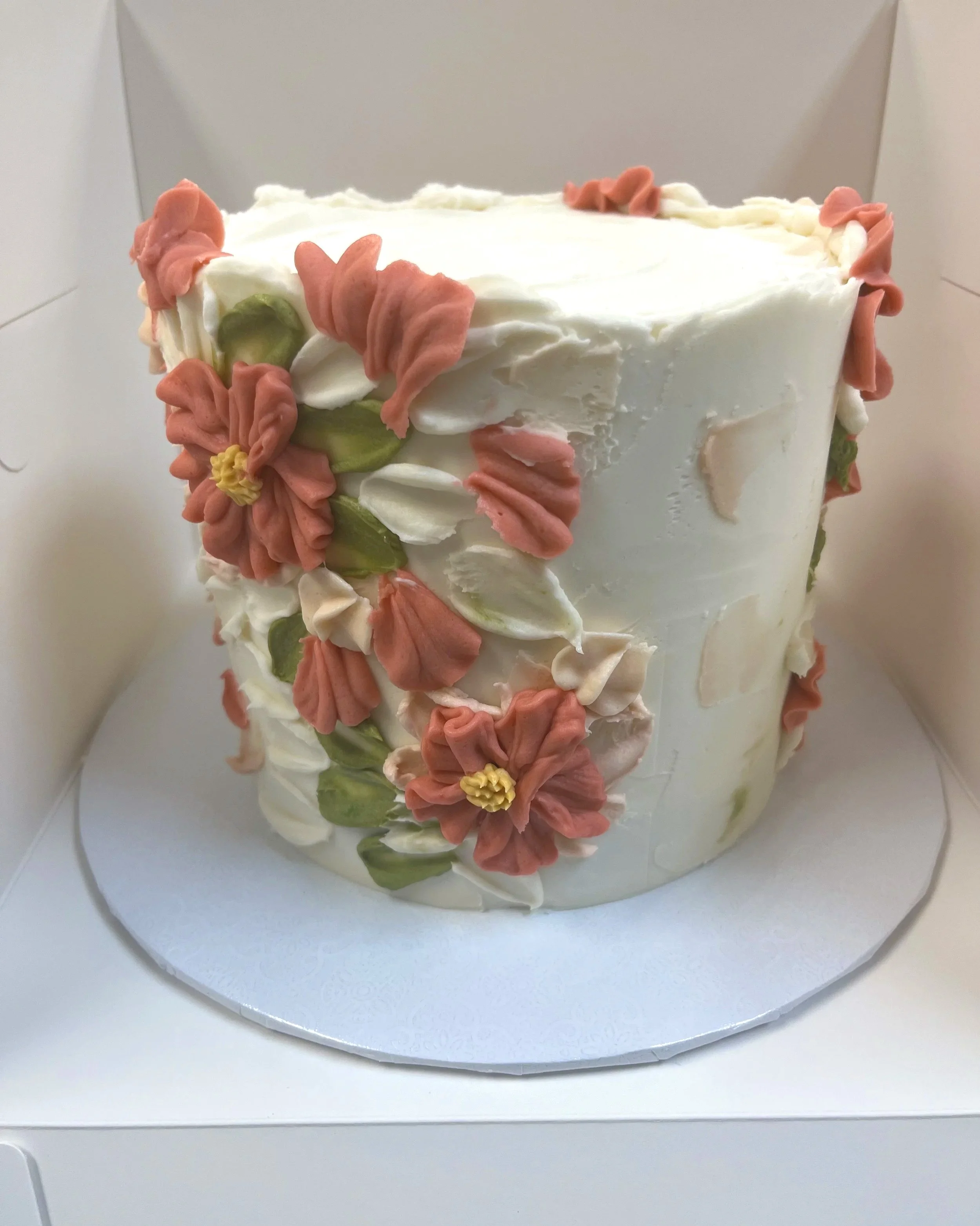 A white frosted birthday cake decorated with pink, white, and green floral piping, placed in a white box.