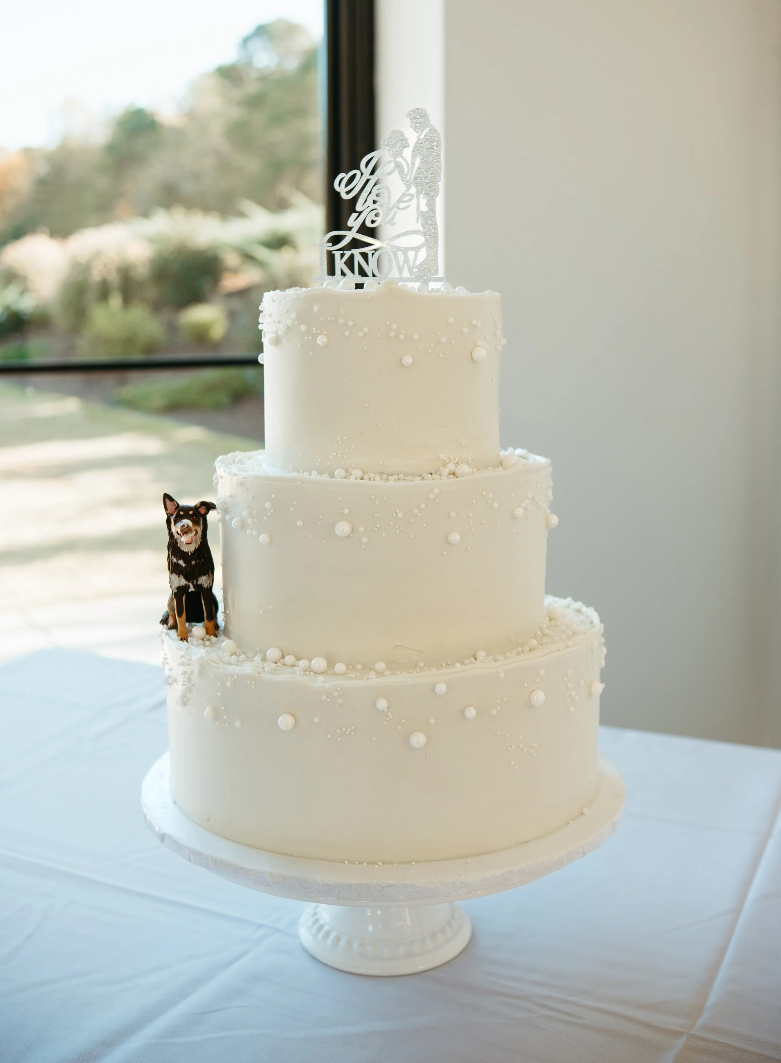 A white three-tier wedding cake decorated with small white pearls and a cake topper featuring a man and woman holding hands, with the words 'Love I Know.' A small dog figurine is attached to the side of the cake.