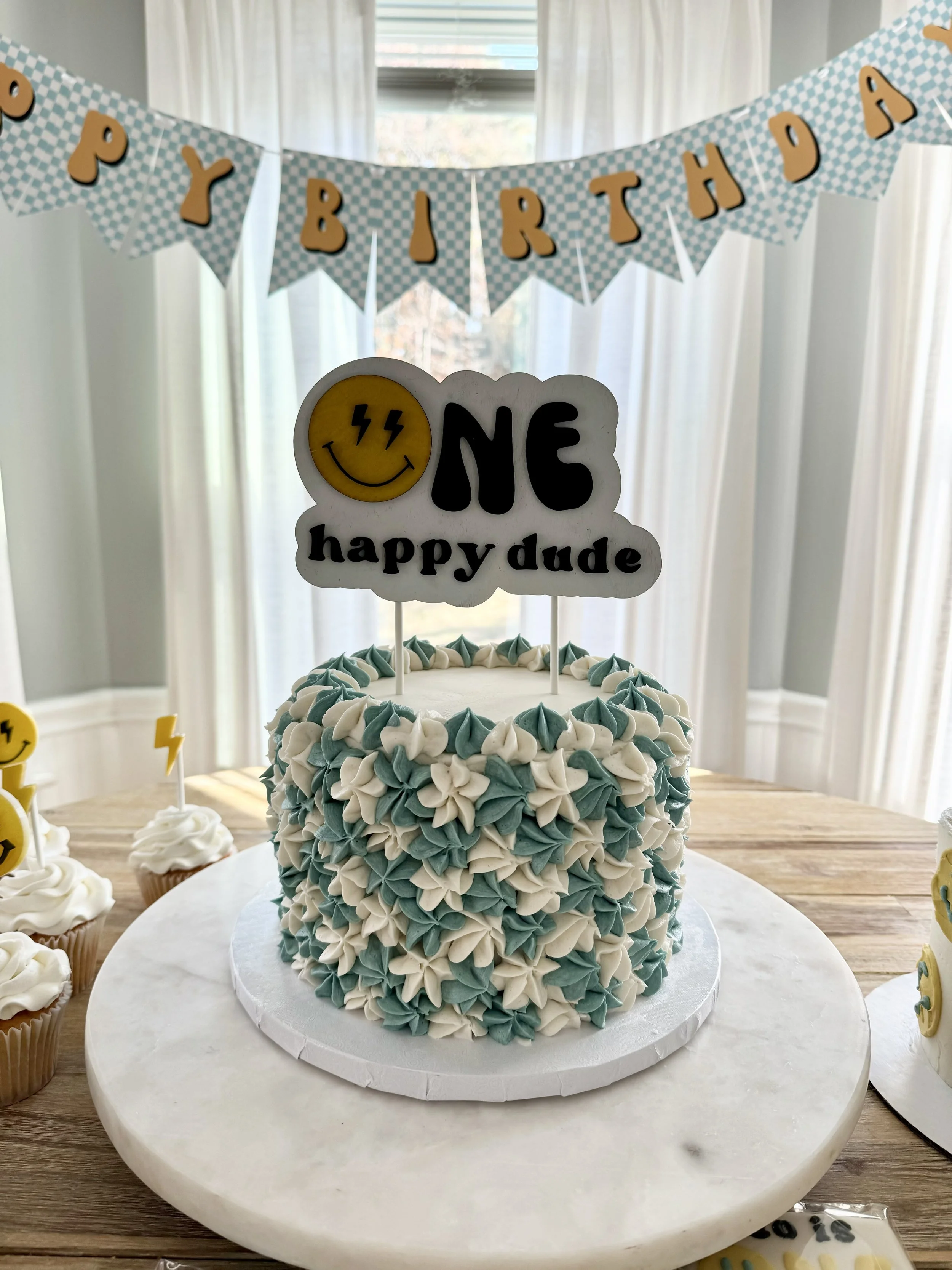 A birthday cake decorated with blue and white frosting flowers, topped with a sign that says 'ONE happy dude' with a smiley face, and a 'Happy Birthday' banner hanging above.