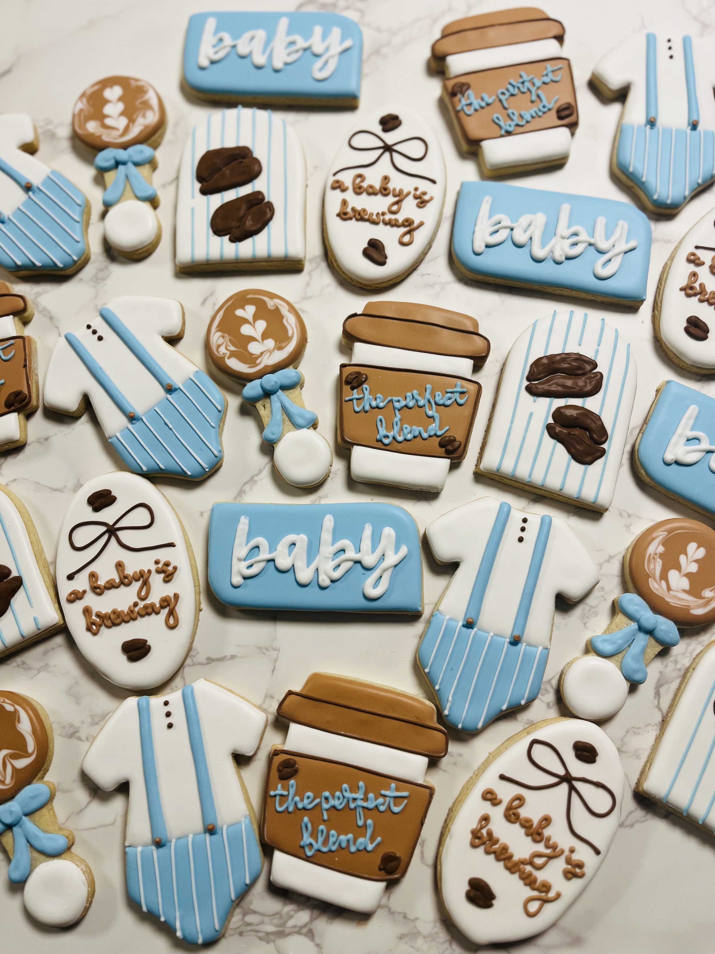 Assorted baby-themed cookies with pastel blue, white, brown, and black icing, decorated with words like "baby" and "the perfect blend," and images of diapers, coffee cups, and baby bottles on a white marble surface.