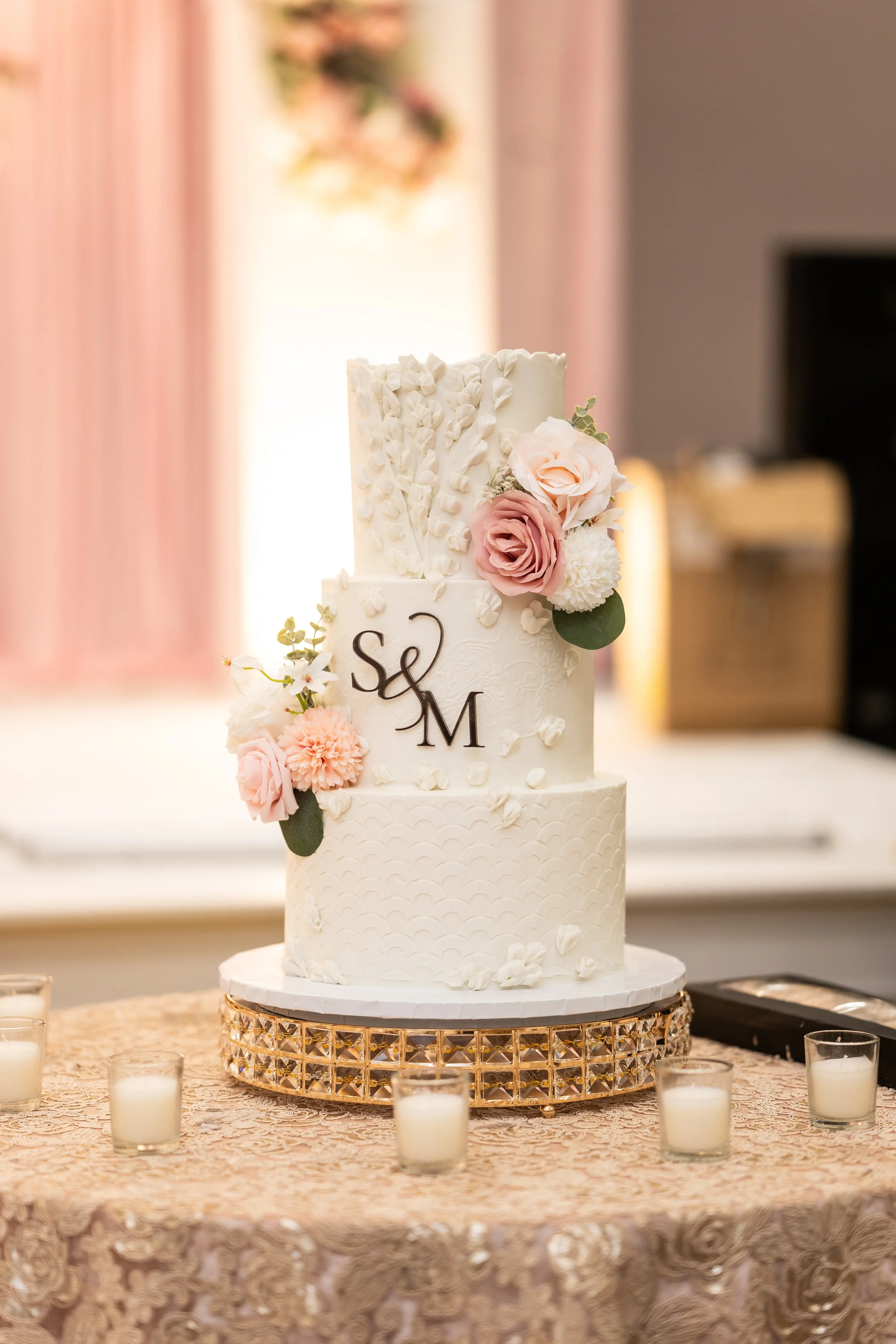 A three-tiered white wedding cake decorated with pink and white roses, placed on a gold woven cake stand on a lace-covered table surrounded by white candles in glass holders.