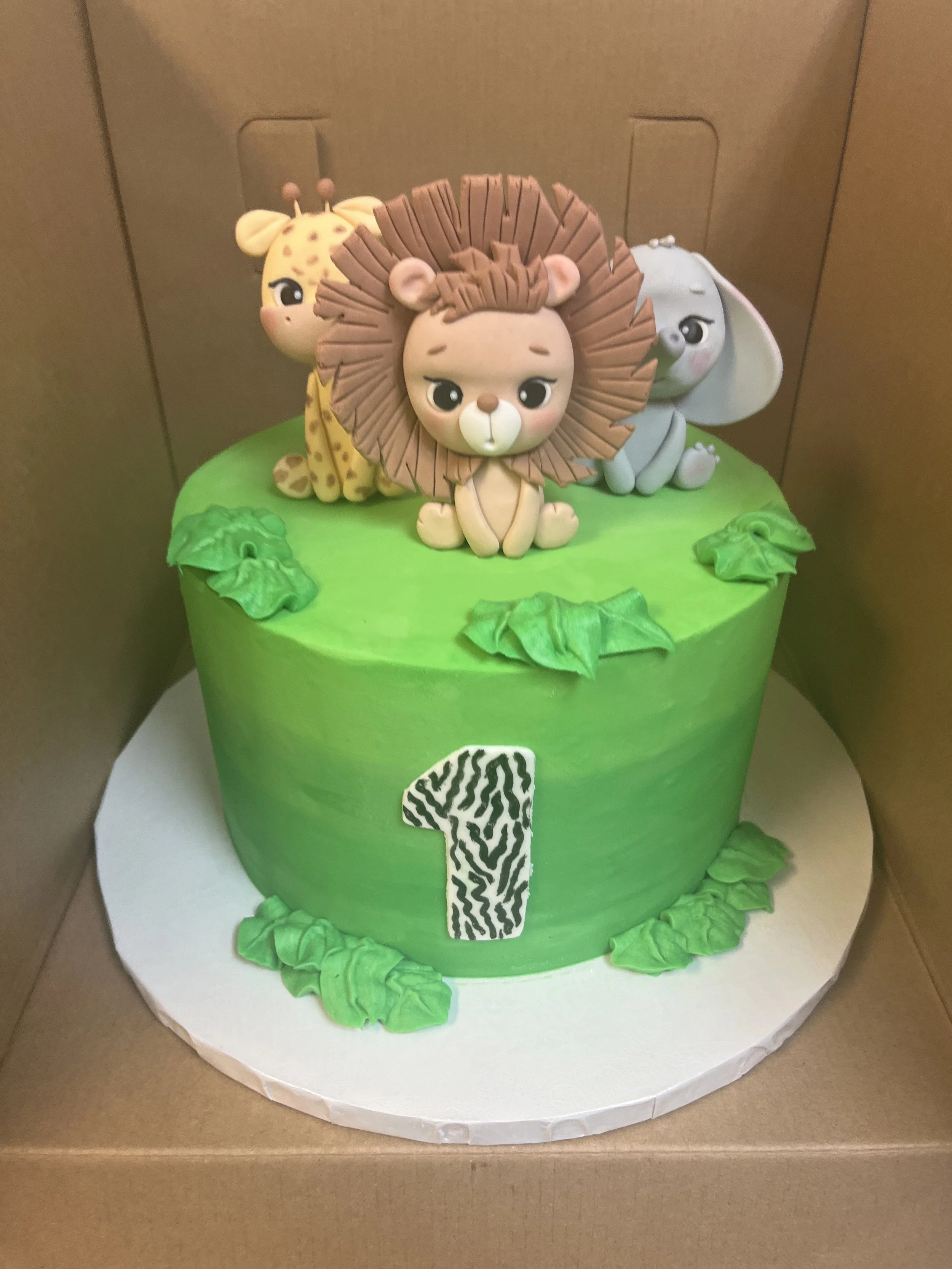 A green jungle-themed birthday cake decorated with fondant animal figurines of a lion, giraffe, and elephant, and a zebra-print number 1 on the side.