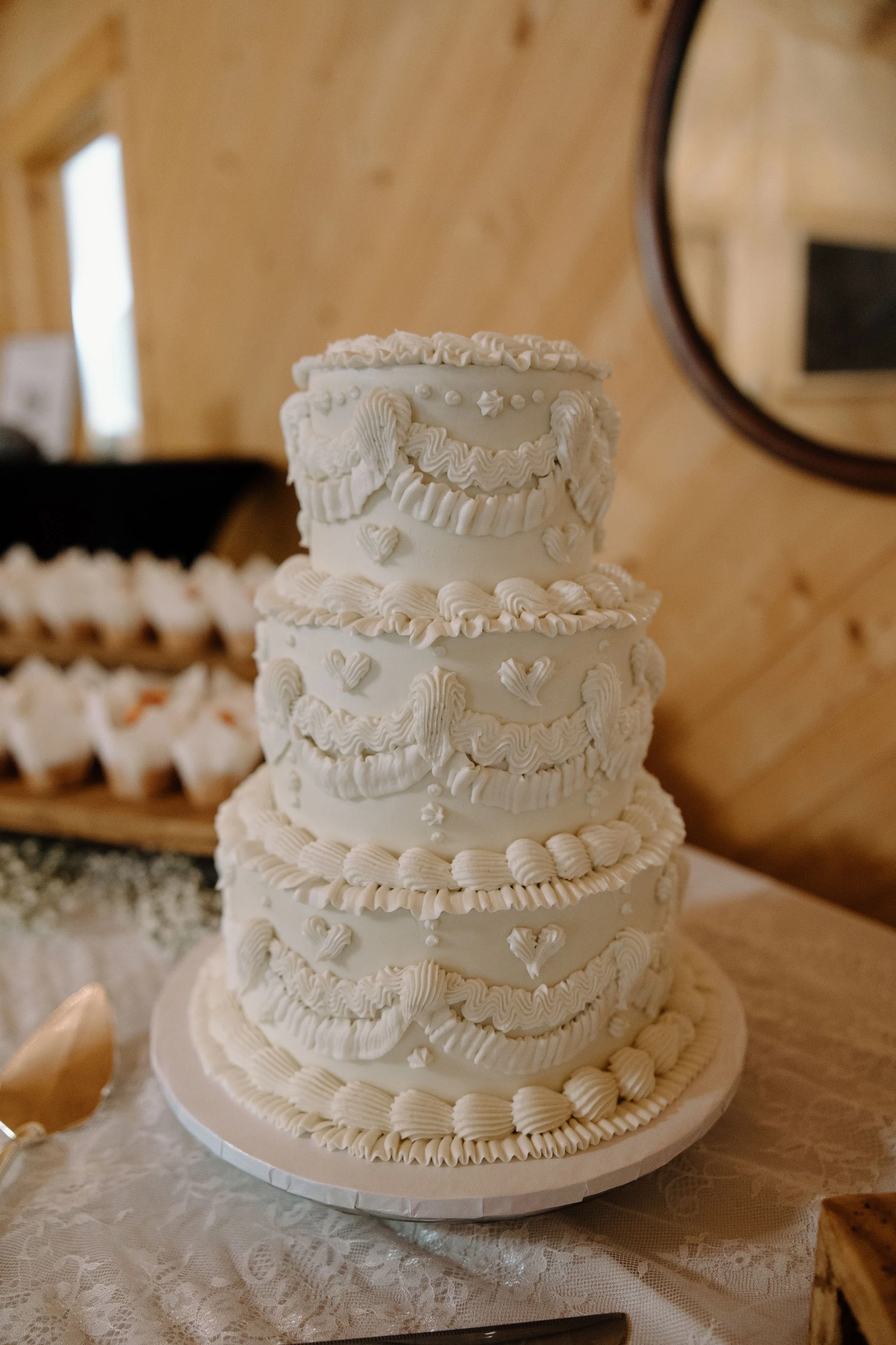 A tall, multi-tiered white wedding cake decorated with intricate icing designs, including swirls, rosettes, and heart shapes, placed on a table with a lace tablecloth.