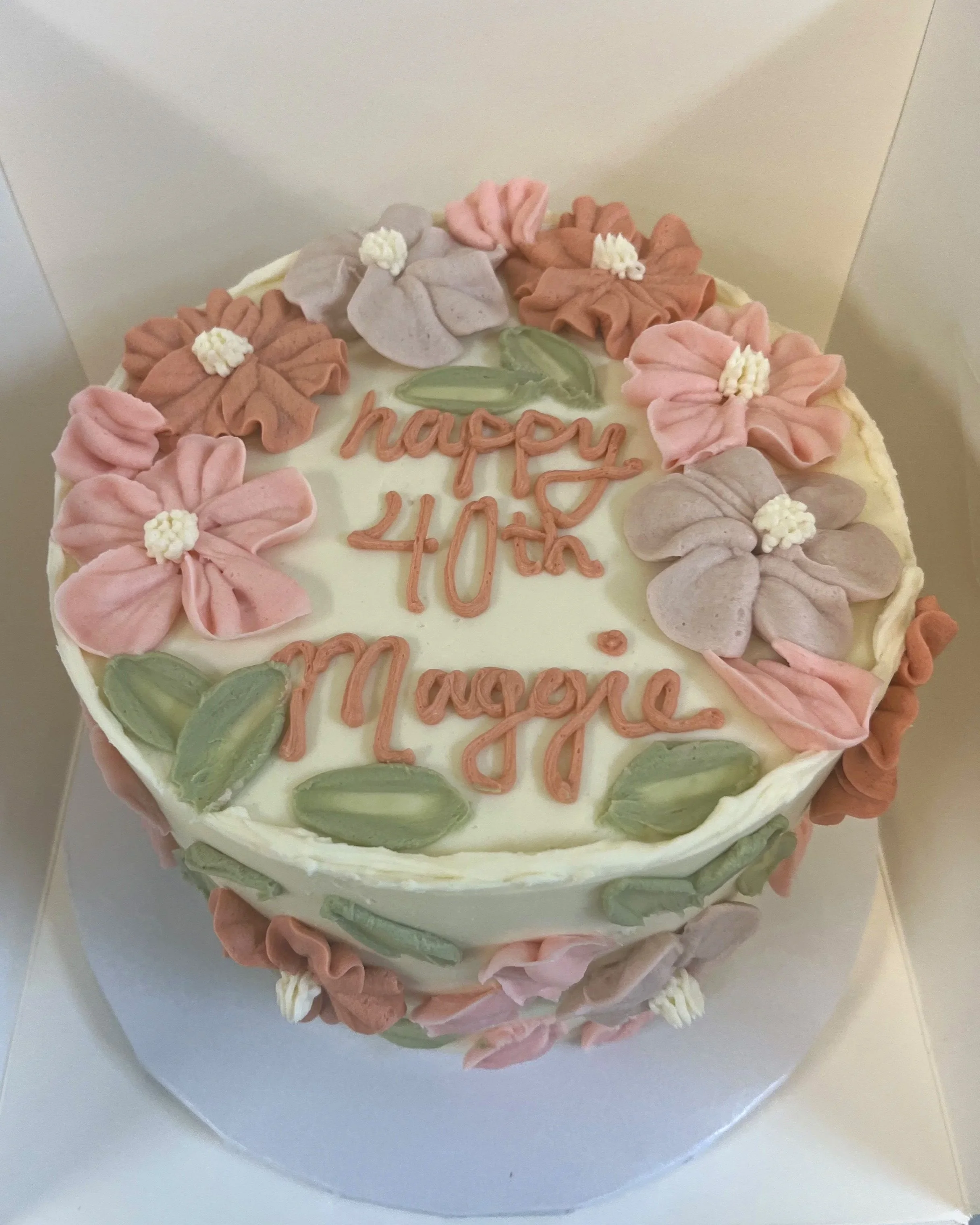 Round cake decorated with pastel-colored flowers and leaves, with a message reading 'happy 40th in progress' in pink icing.