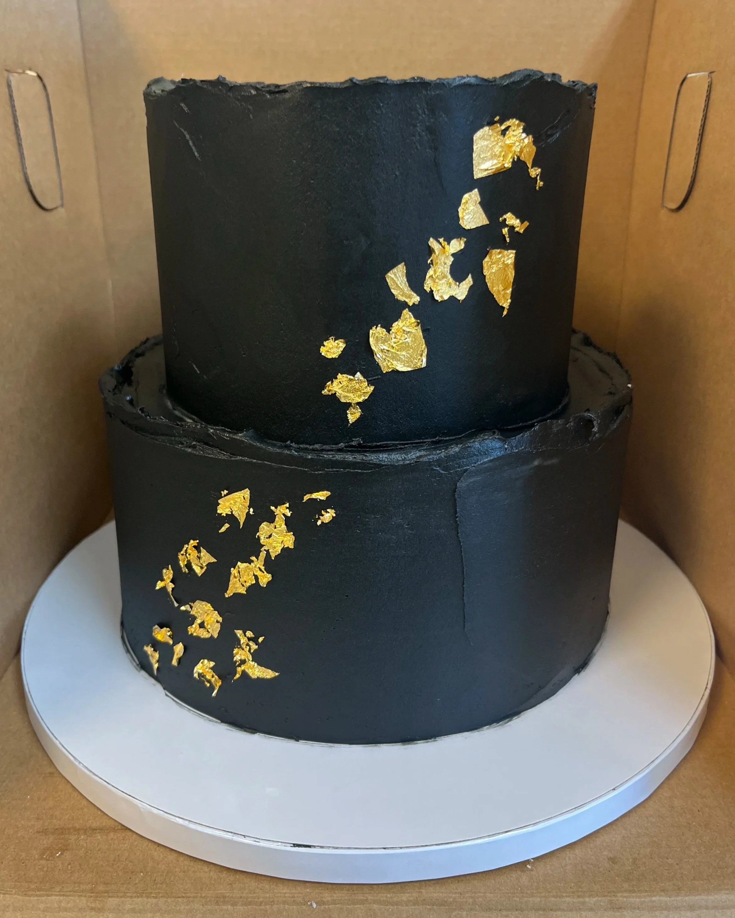 Two-tier black cake decorated with gold leaf accents, placed in a cardboard box on a white cake board.