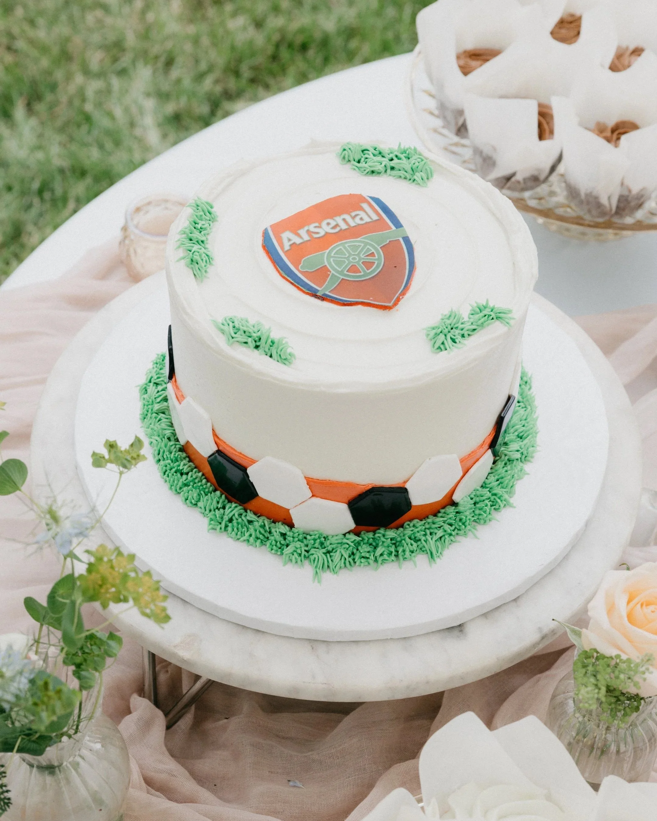 A soccer-themed birthday cake with an Arsenal logo on top, decorated with green frosting and black and white soccer ball pattern around the sides, on a white marble table.