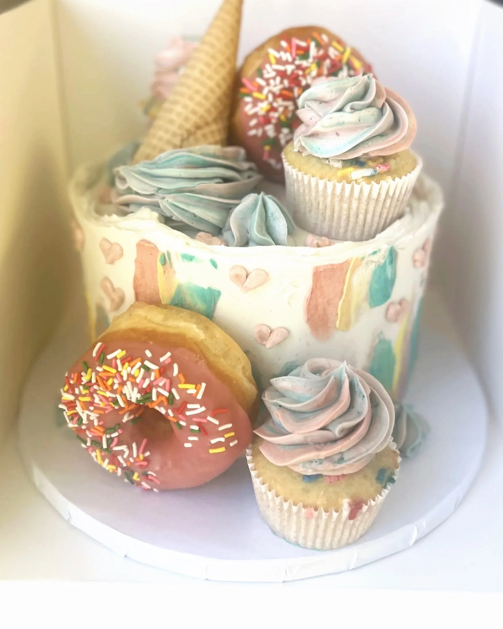 A decorative cake with pastel-colored frosting, sprinkles, cupcakes, donuts, and a decorative cone on top.
