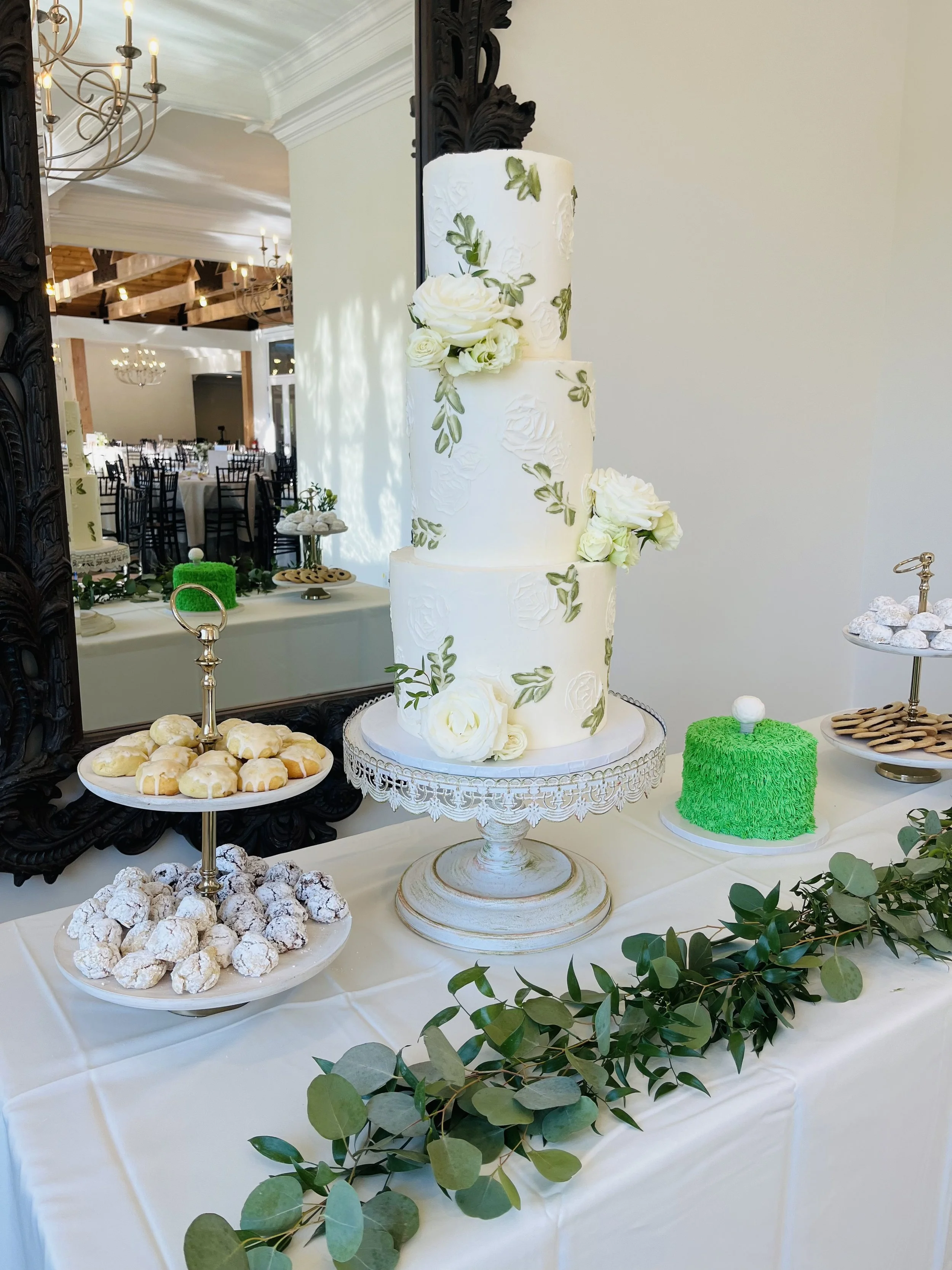 A three-tier white wedding cake decorated with white roses and green leaves, set on a vintage-style cake stand. The table has assorted desserts including powdered sugar-covered cookies and a bright green cake with textured frosting. Greenery runs along the front of the table, and a large mirror reflects the elegant reception hall in the background.