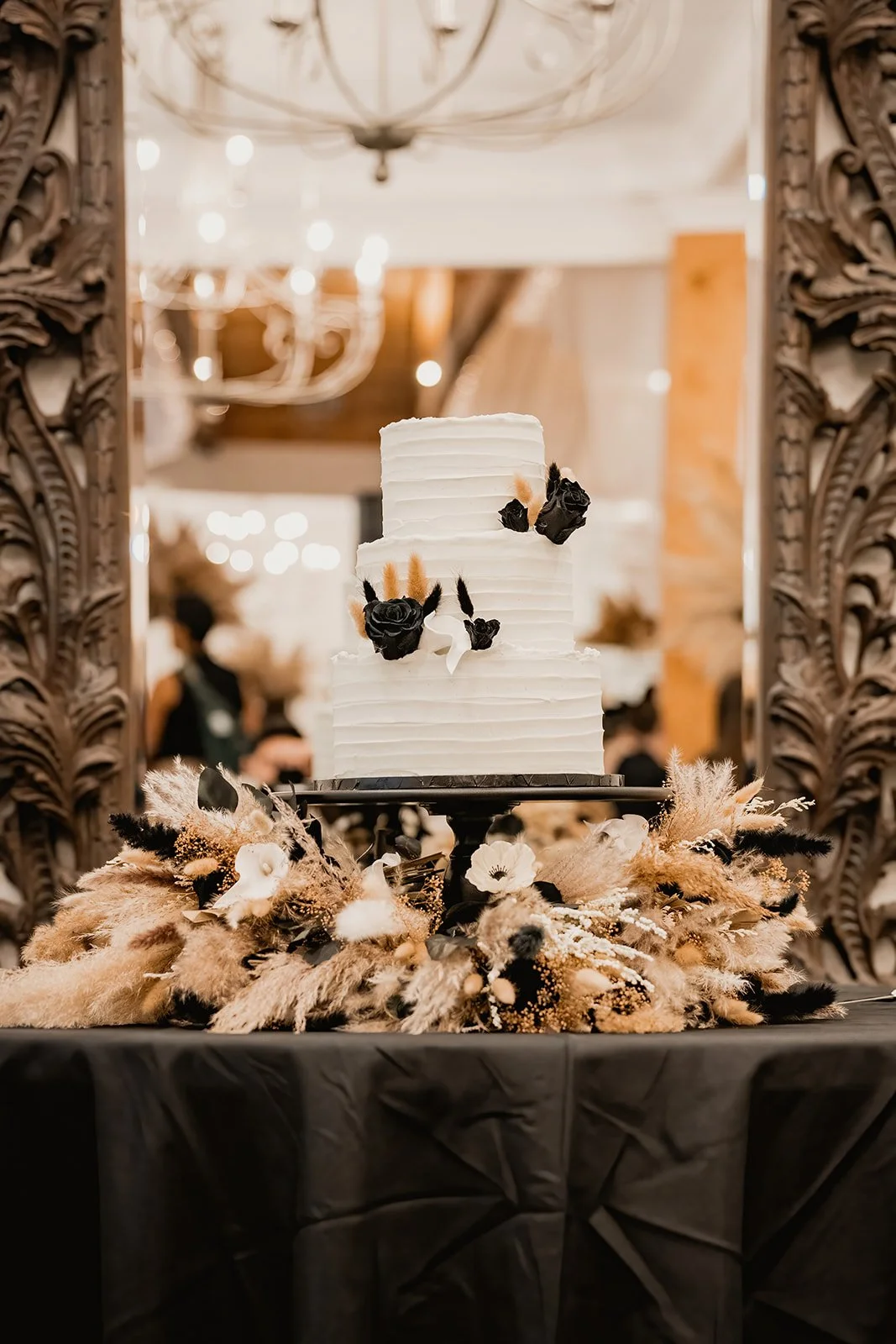 A tall, three-tiered white wedding cake decorated with black roses and small black accents, surrounded by beige and black dried floral arrangements on a black table, with ornate framing and warm lighting in the background.