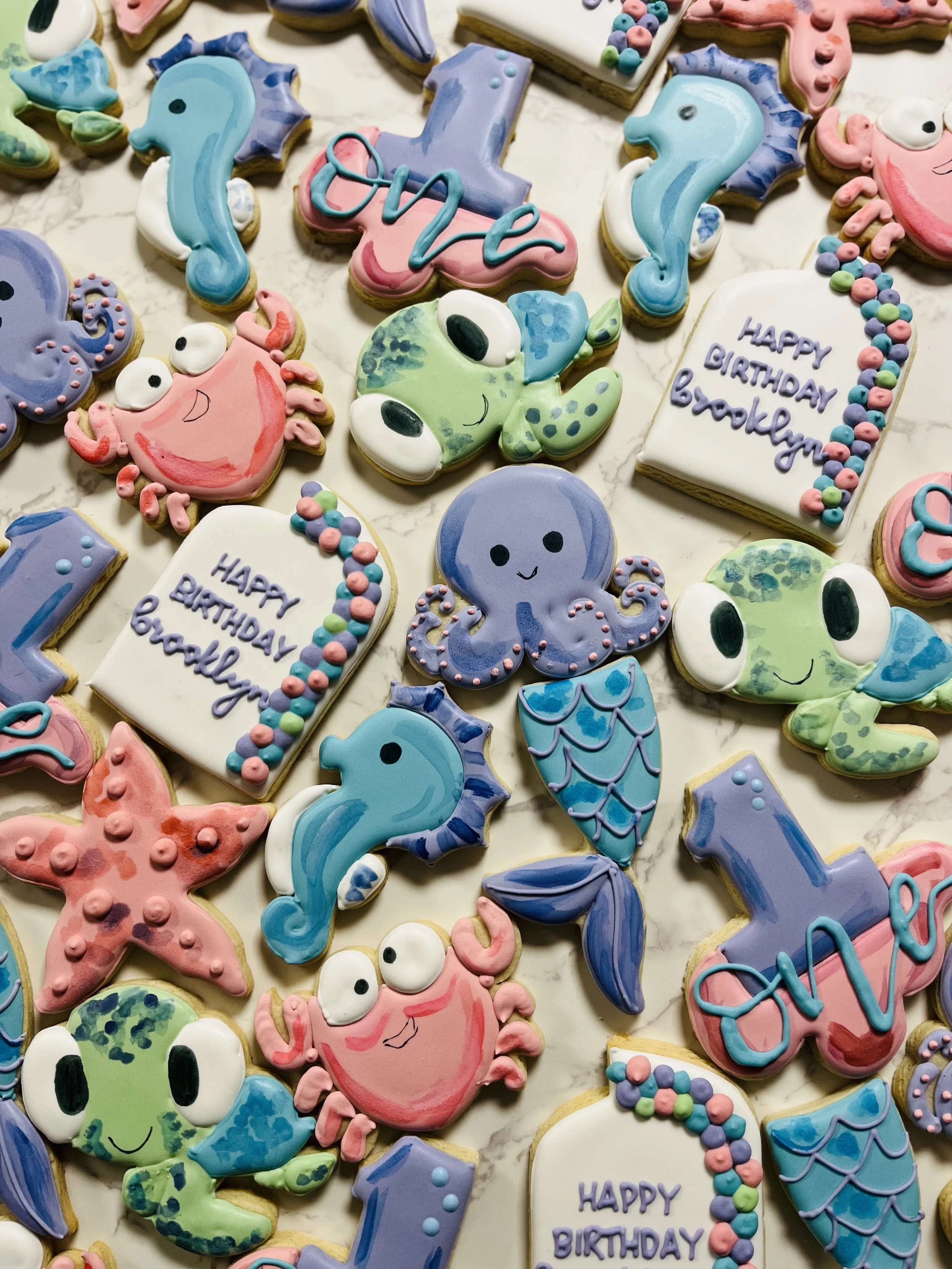 Assorted colorful birthday cookies decorated with ocean-themed designs such as seahorses, crabs, fish, and whales, along with occasion specific cookies with messages like 'Happy Birthday Brooklyn'.