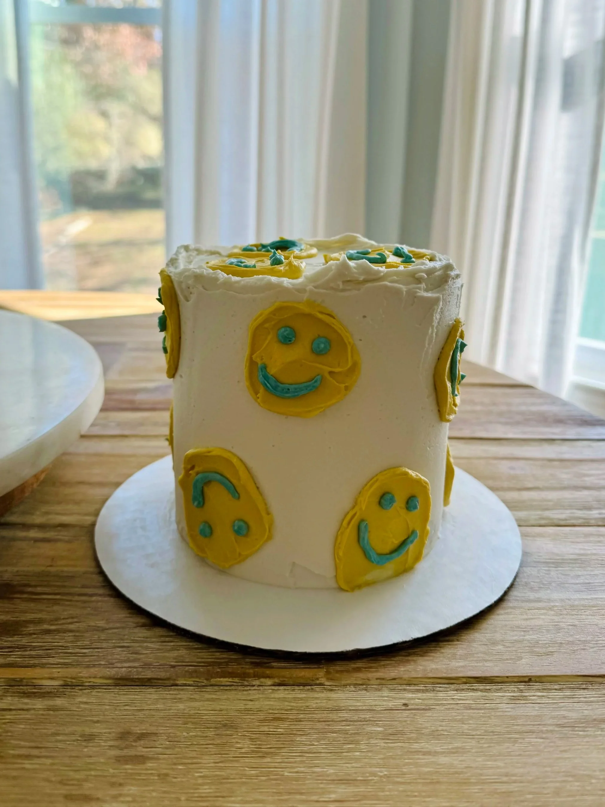 A white cake decorated with yellow and blue smiley face designs, placed on a wooden table near a window.