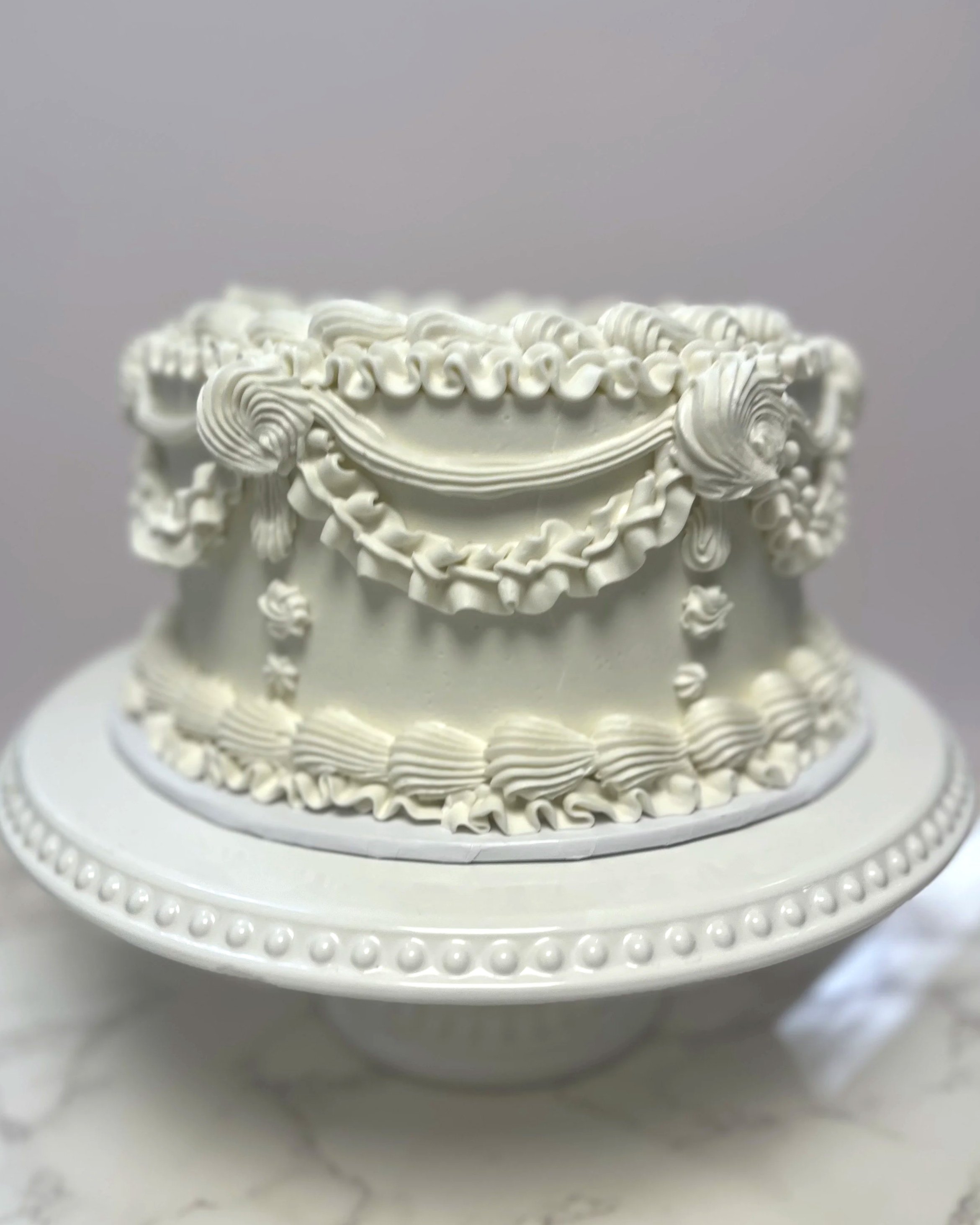 White decorative cake with ornate piping and design, placed on a white cake stand.