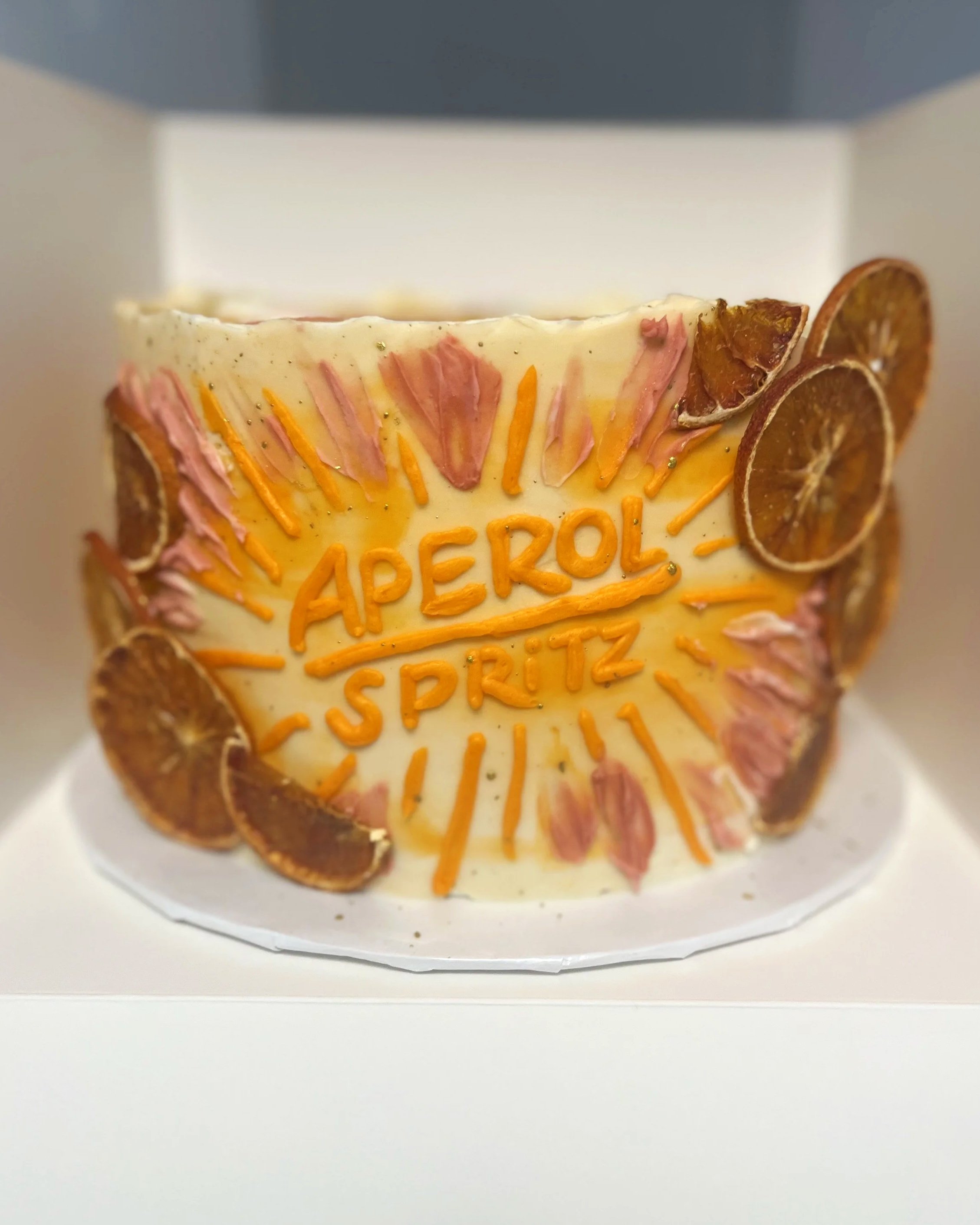 Celebration cake with a white frosting exterior, decorated with dried orange slices, pink and orange icing accents, and the words 'Aperol Spritz' written in orange icing.