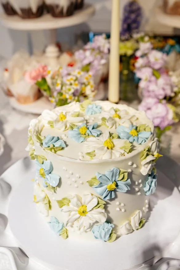Decorated cake with white, blue, and yellow floral icing on a white cake stand, with a yellow candle on top, surrounded by flower arrangements.
