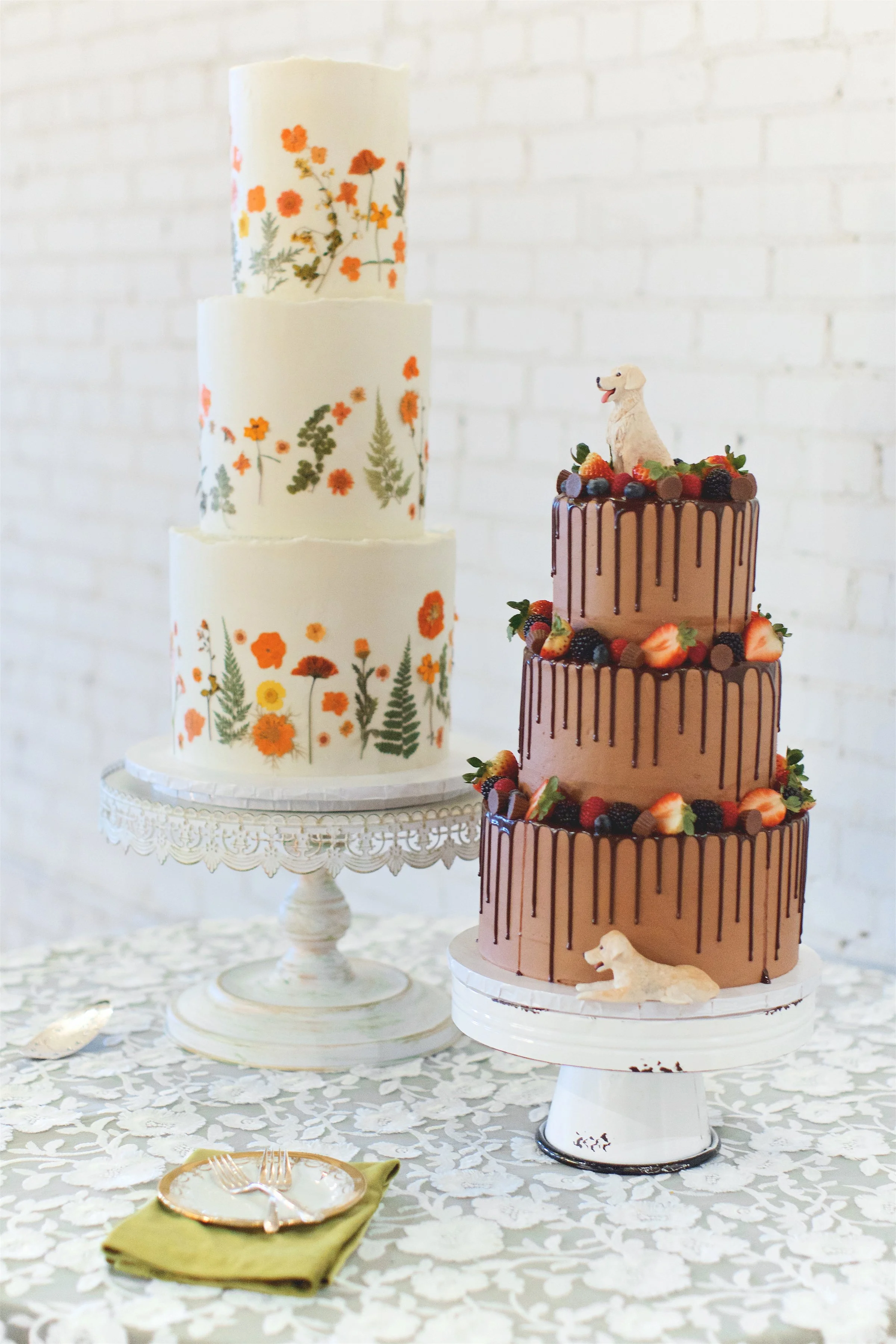 Two decorated cakes on cake stands. The taller cake on the left has watercolor floral patterns. The shorter, three-tiered chocolate cake on the right is topped with berries and dog figurines.