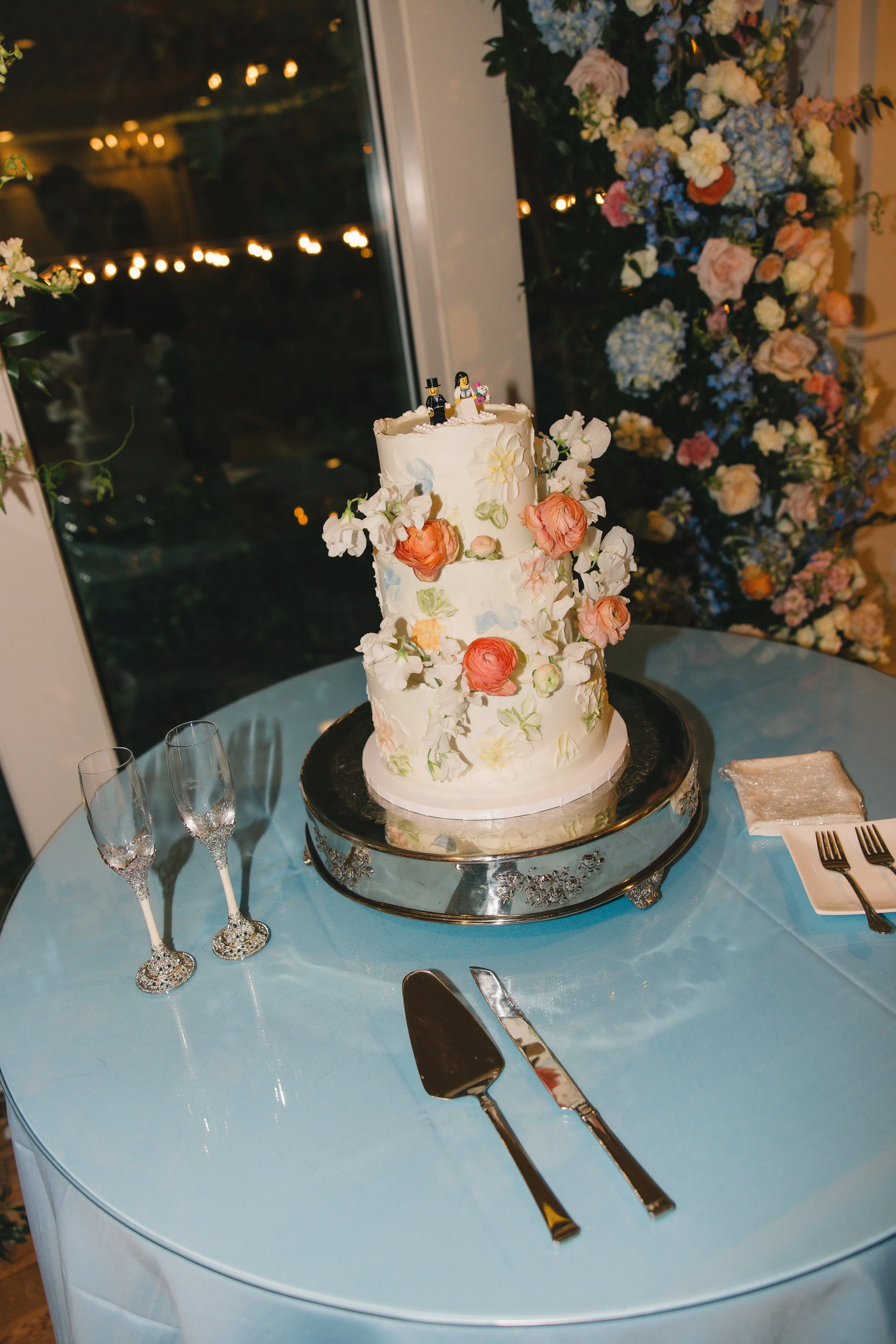 A wedding cake with white icing decorated with pastel-colored flowers, topped with wedding figurines, placed on a silver tray on a blue tablecloth, with champagne flutes and silverware nearby, and a flower arrangement in the background.