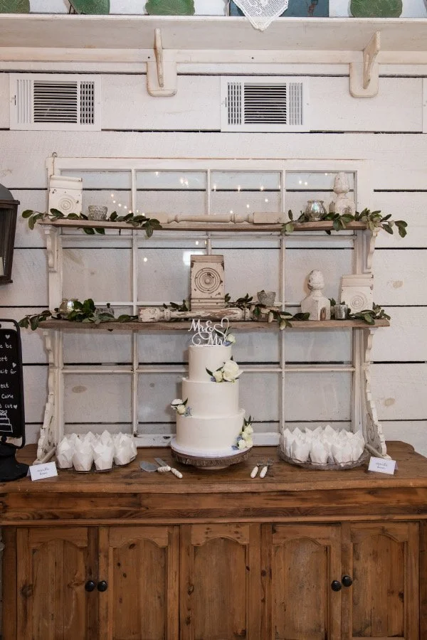 White wedding cake with white flowers on a wooden table, surrounded by white party favor bags, with a rustic white wooden backdrop decorated with greenery and vintage items.
