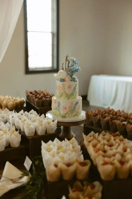A multi-tiered cake decorated with flowers and pastel colors on a table surrounded by various small desserts at a celebration or party.