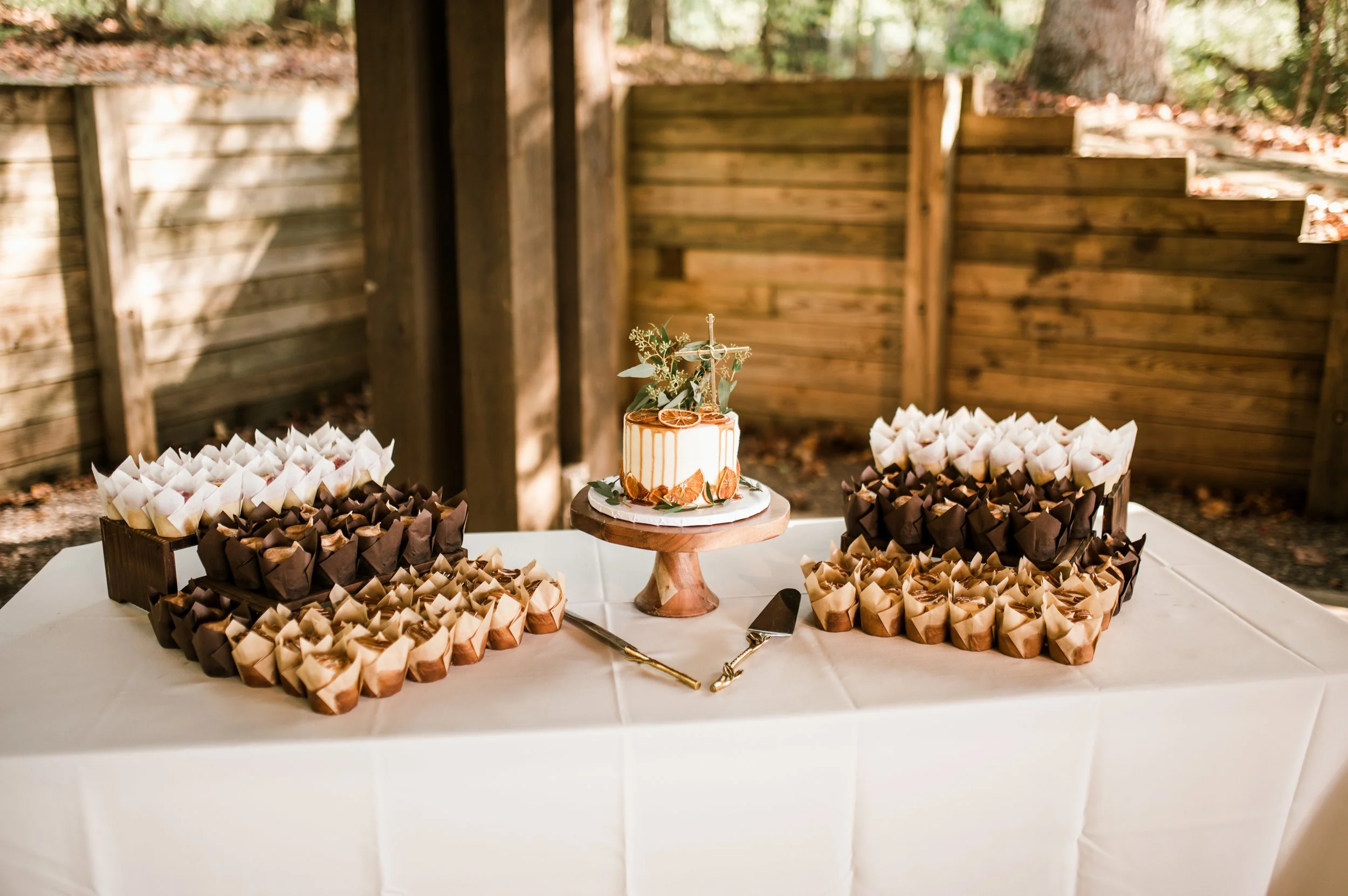 A white table with a small decorated cake on a wooden stand and many small cupcake-like desserts wrapped in white, brown, and tan paper.