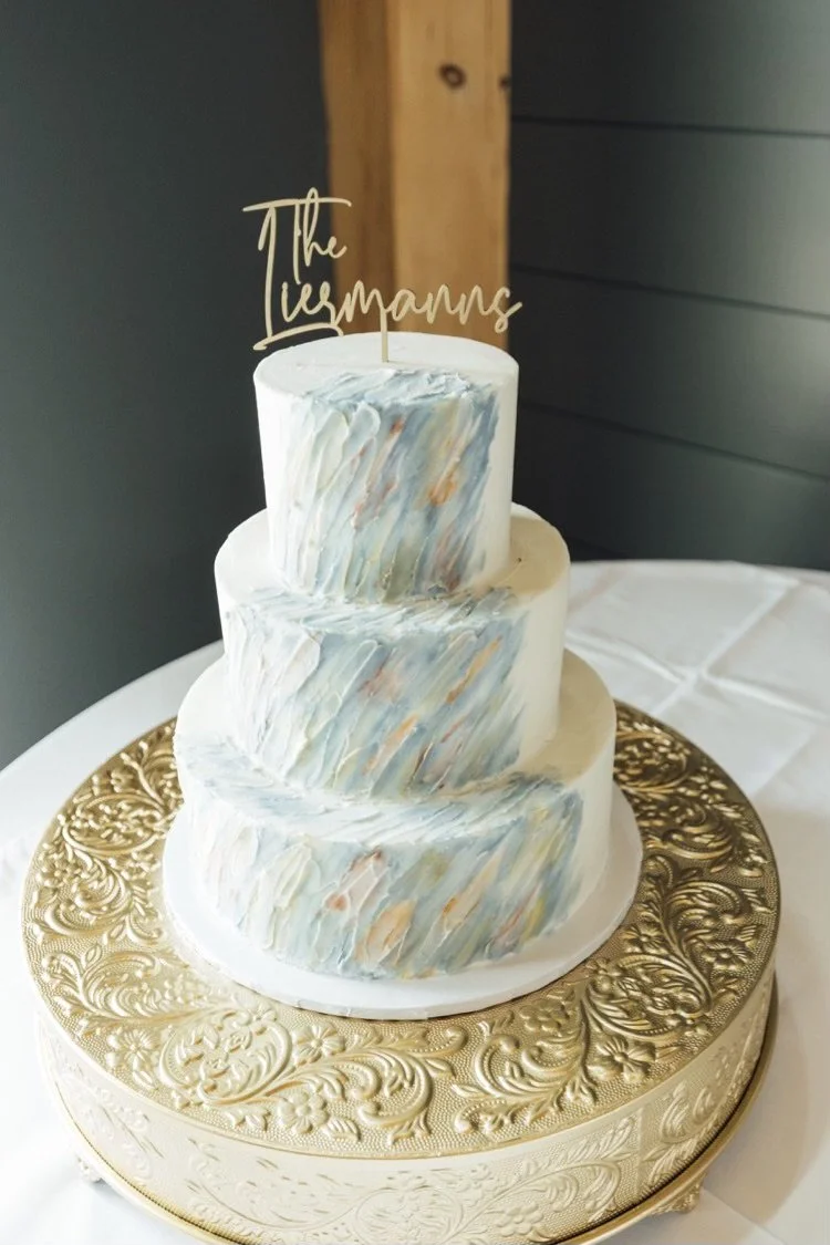 A three-tiered wedding cake with a marbled blue, white, and peach design, placed on a gold decorative cake stand, with a gold acrylic cake topper that reads 'The Ilugmanns'.