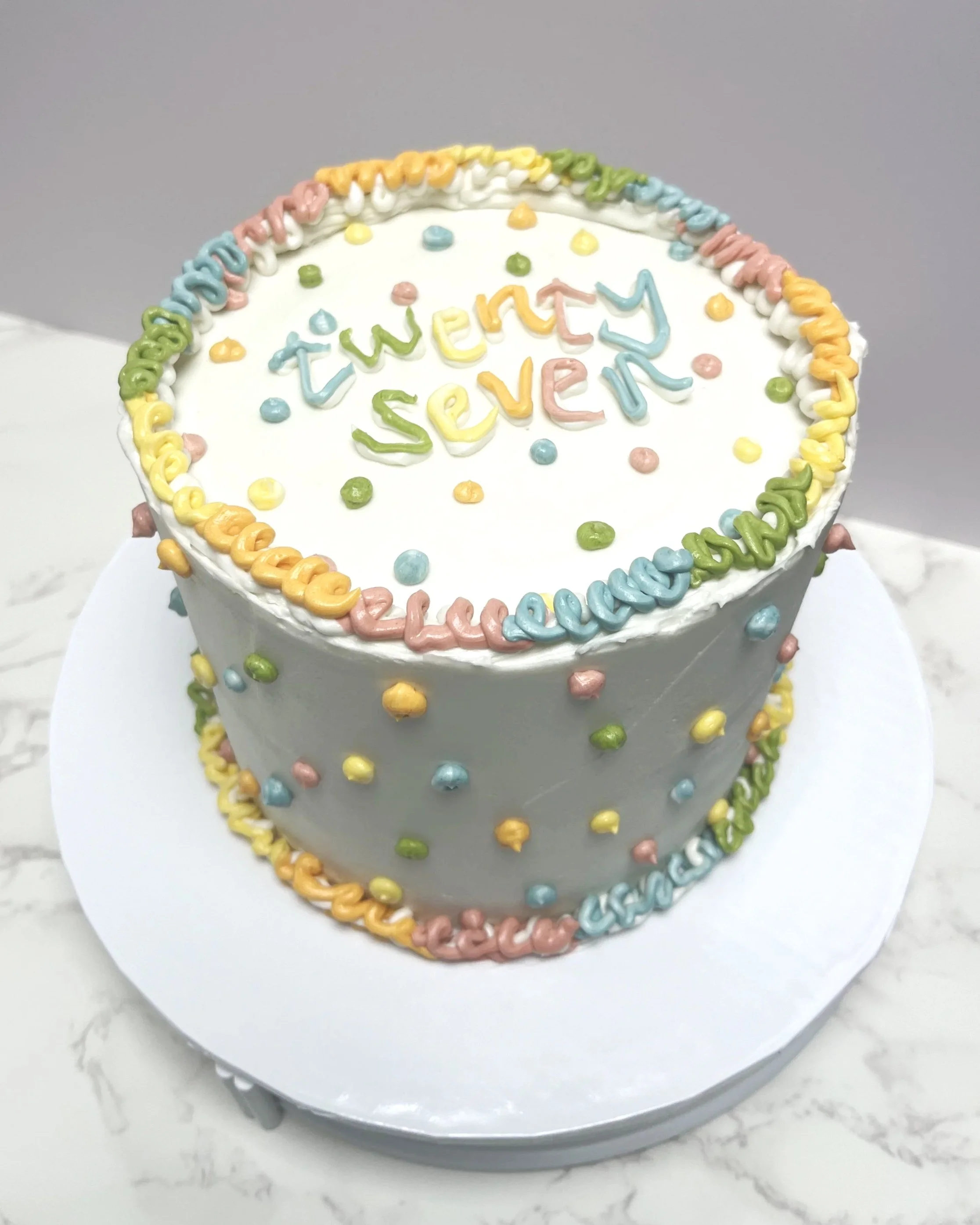 Colorful birthday cake with pastel sprinkles and the words 'twenty seven' written on top.