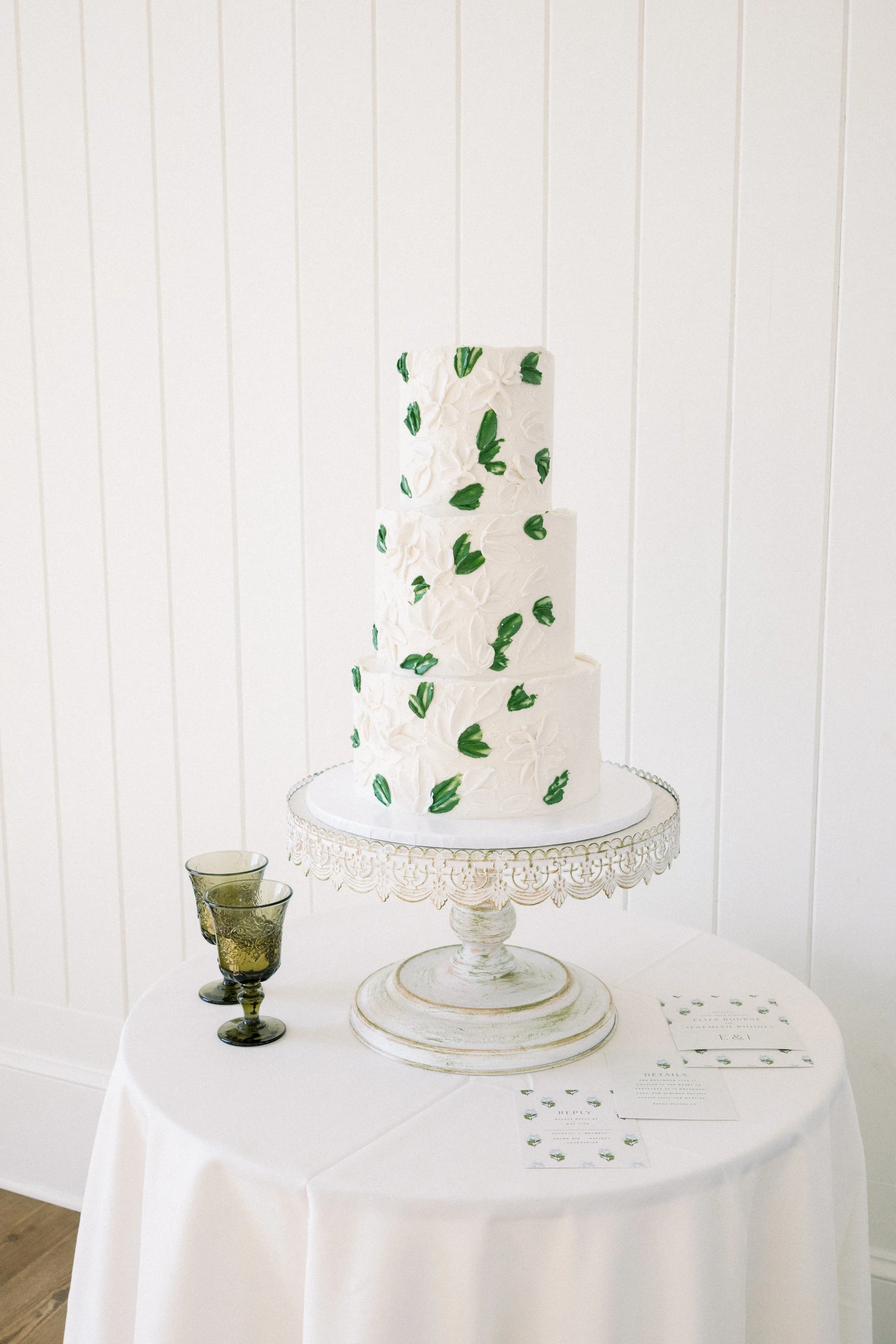 A multi-tiered white wedding cake decorated with white and green floral designs, placed on a decorative cake stand on a white table. There are two glasses with a dark liquid and some printed papers or programs on the table.