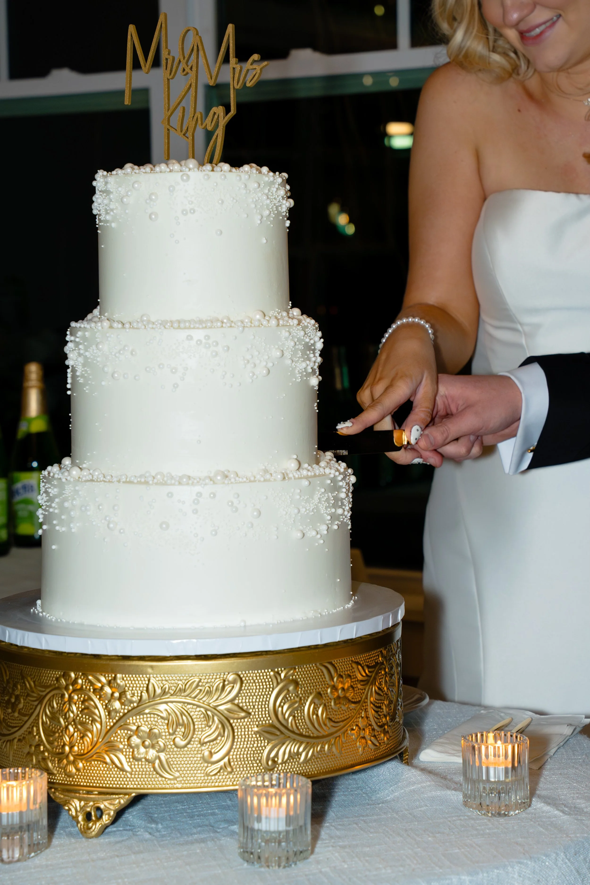 A three-tier white wedding cake decorated with white pearls and beads, topped with a gold "Mrs. & Mr's" cake topper, on a gold ornate cake stand, with candles and tableware nearby.