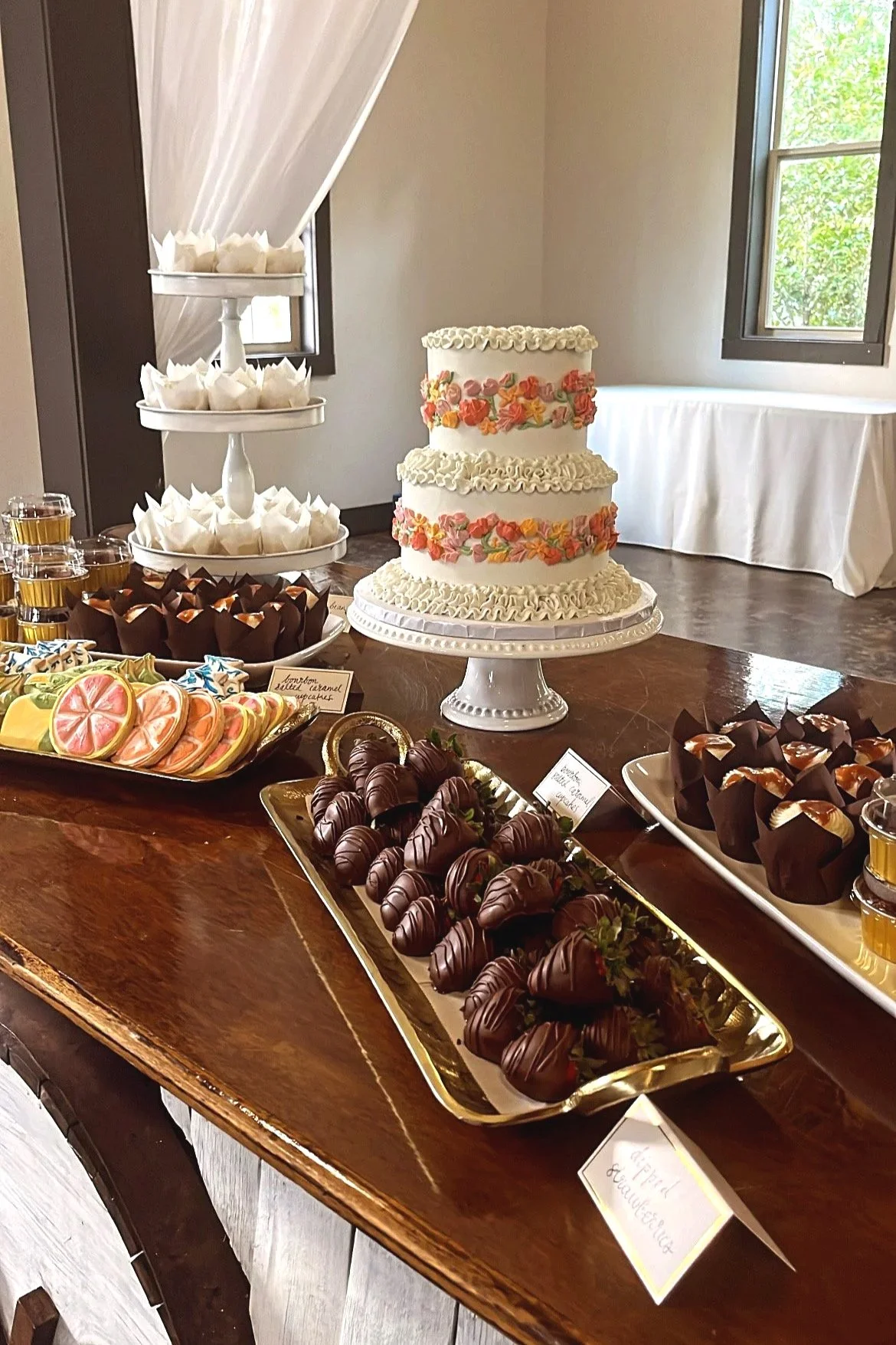 A three-tiered wedding cake decorated with flowers in pink, orange, and yellow rosettes. The cake is on a white cake stand on a wooden table. Surrounding the cake are various chocolates, candies, and desserts on platters and trays.