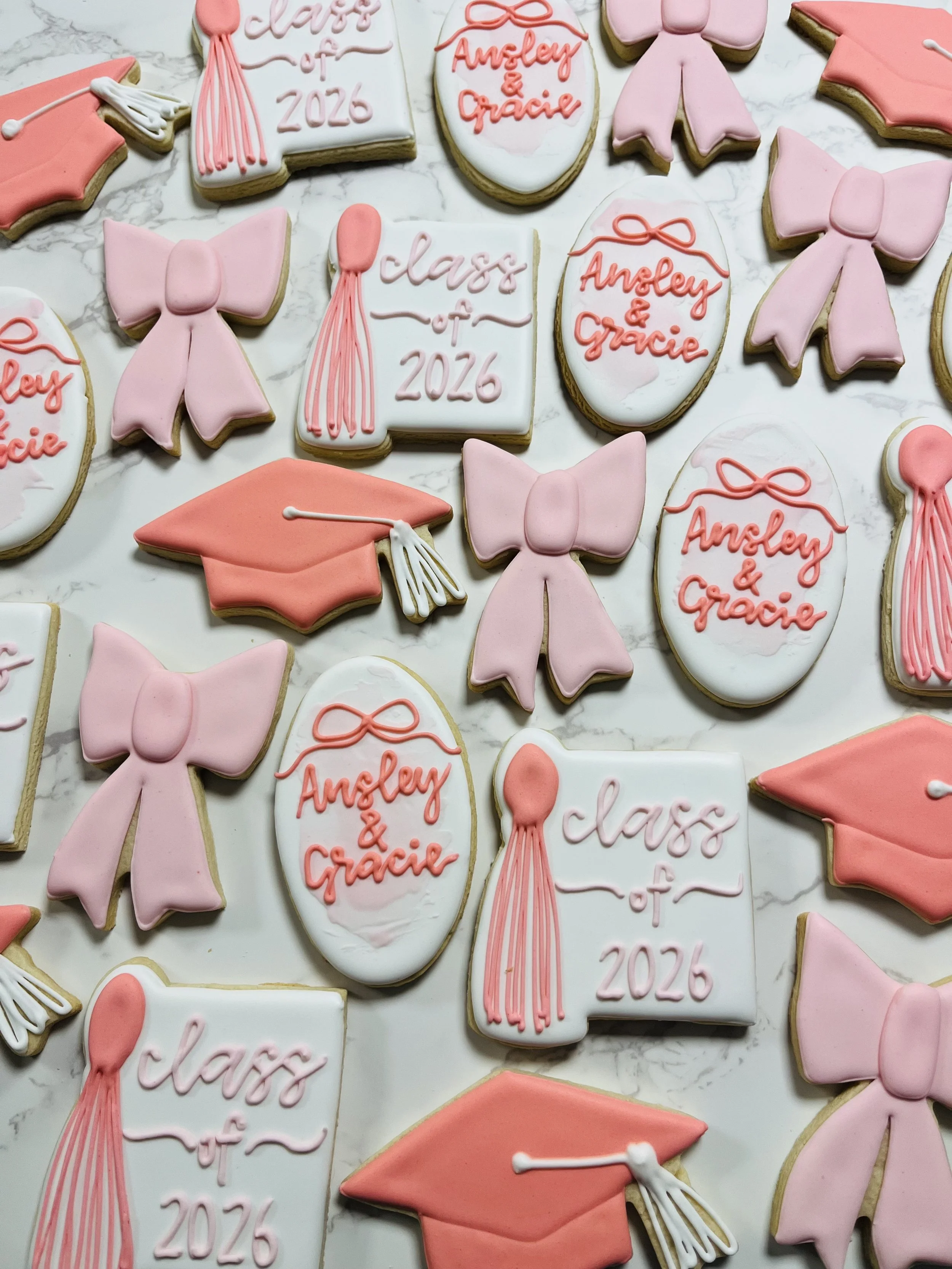 Decorated cookies for a graduation celebration, featuring pink and white icing with messages such as "Class of 2026" and "Angley & Gracie," along with cookies shaped like graduation caps, bows, and diploma scrolls.