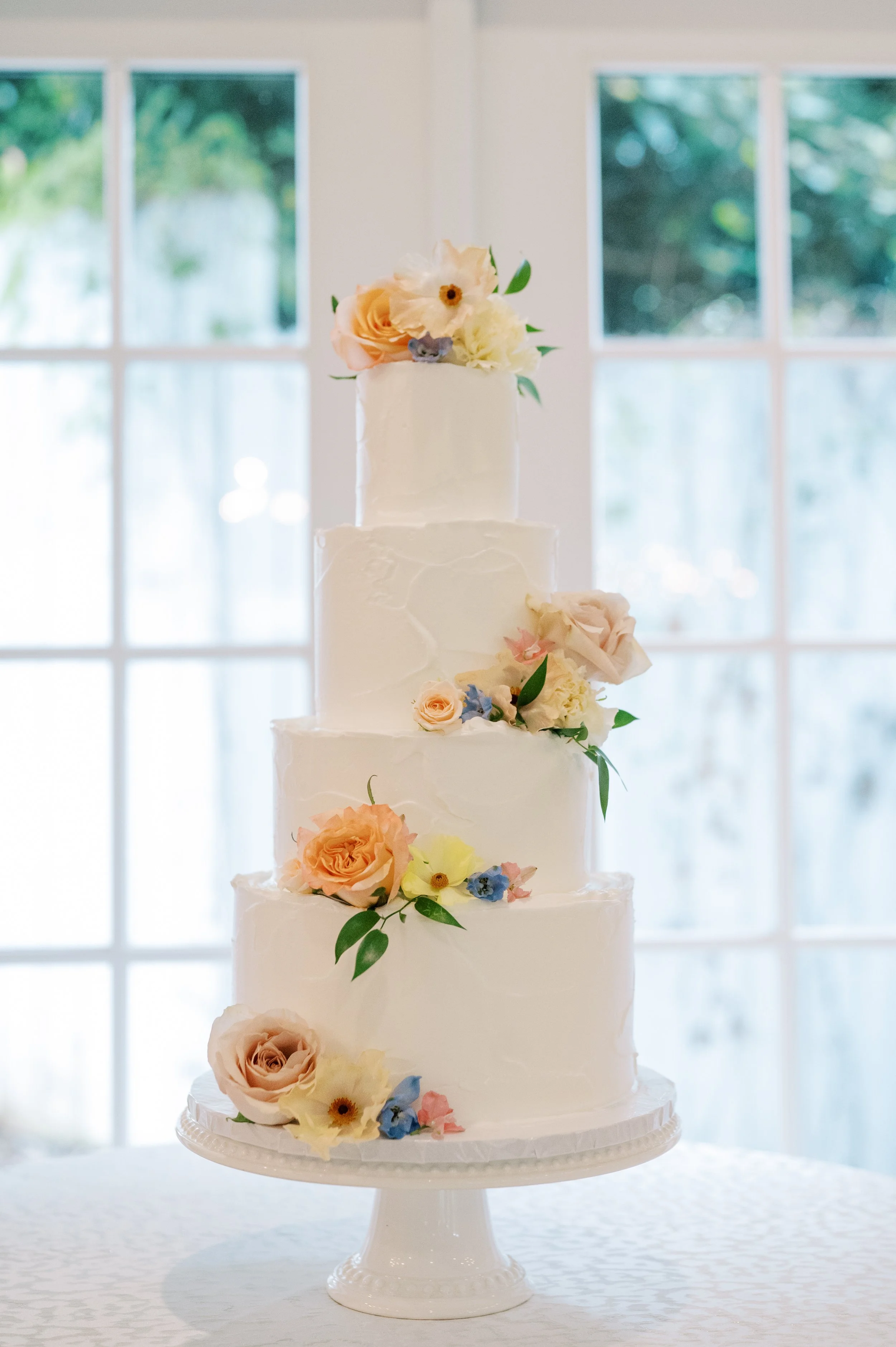 A four-tier white wedding cake decorated with pastel-colored roses and flowers, displayed on a white cake stand in front of a window.