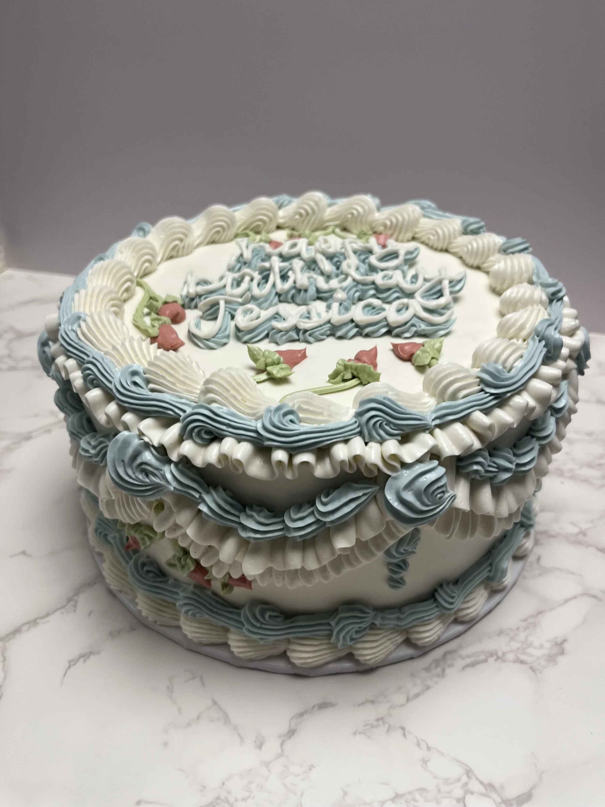 A round birthday cake decorated with white, blue, and green piping, with pink floral accents, and a message that says 'Happy Birthday Jacqueline' on top.