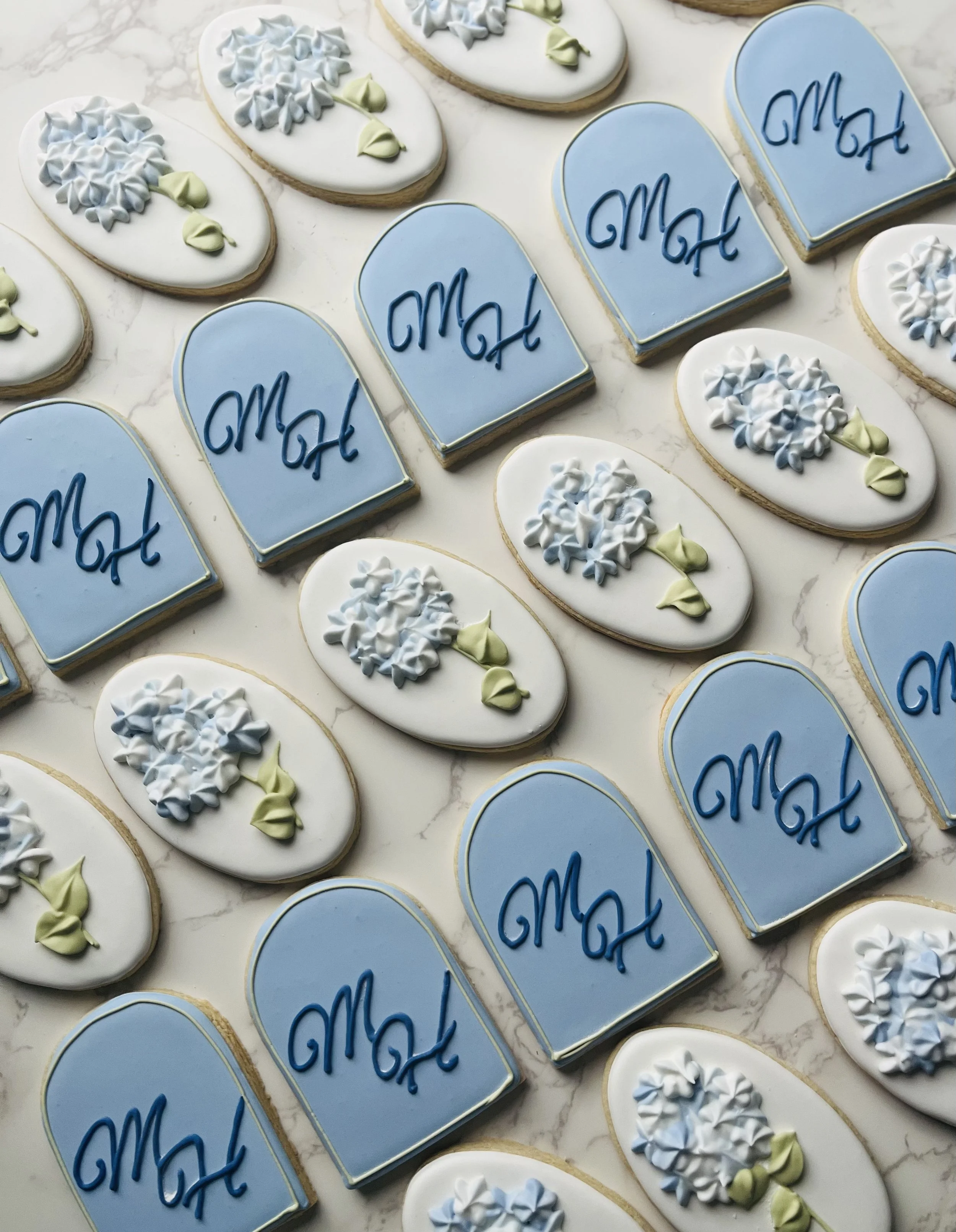 Various decorated sugar cookies on a marble surface, with shapes including hearts, ovals, and arches. The cookies have white and blue icing, with some featuring floral patterns and others with the word "Mom" written in dark blue icing.